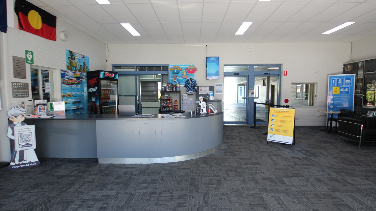 The automatic glass sliding doors at Michael Wendon Aquatic Leisure Centre can be easily distinguished by their white painted edges and grey walls. White square LED light panels are placed on the ceiling. The name of the venue can be found on both upper sides of the entrance doors as well as small and large marketing posters scattered on the glass doors and on the smooth concrete grey wall. The automatic glass sliding doors at Michael Wendon Aquatic Leisure Centre can be easily distinguished by their white painted edges and grey walls. White square LED light panels are placed on the ceiling. The name of the venue can be found on both upper sides of the entrance doors as well as small and large marketing posters scattered on the glass doors and on the smooth concrete grey wall.