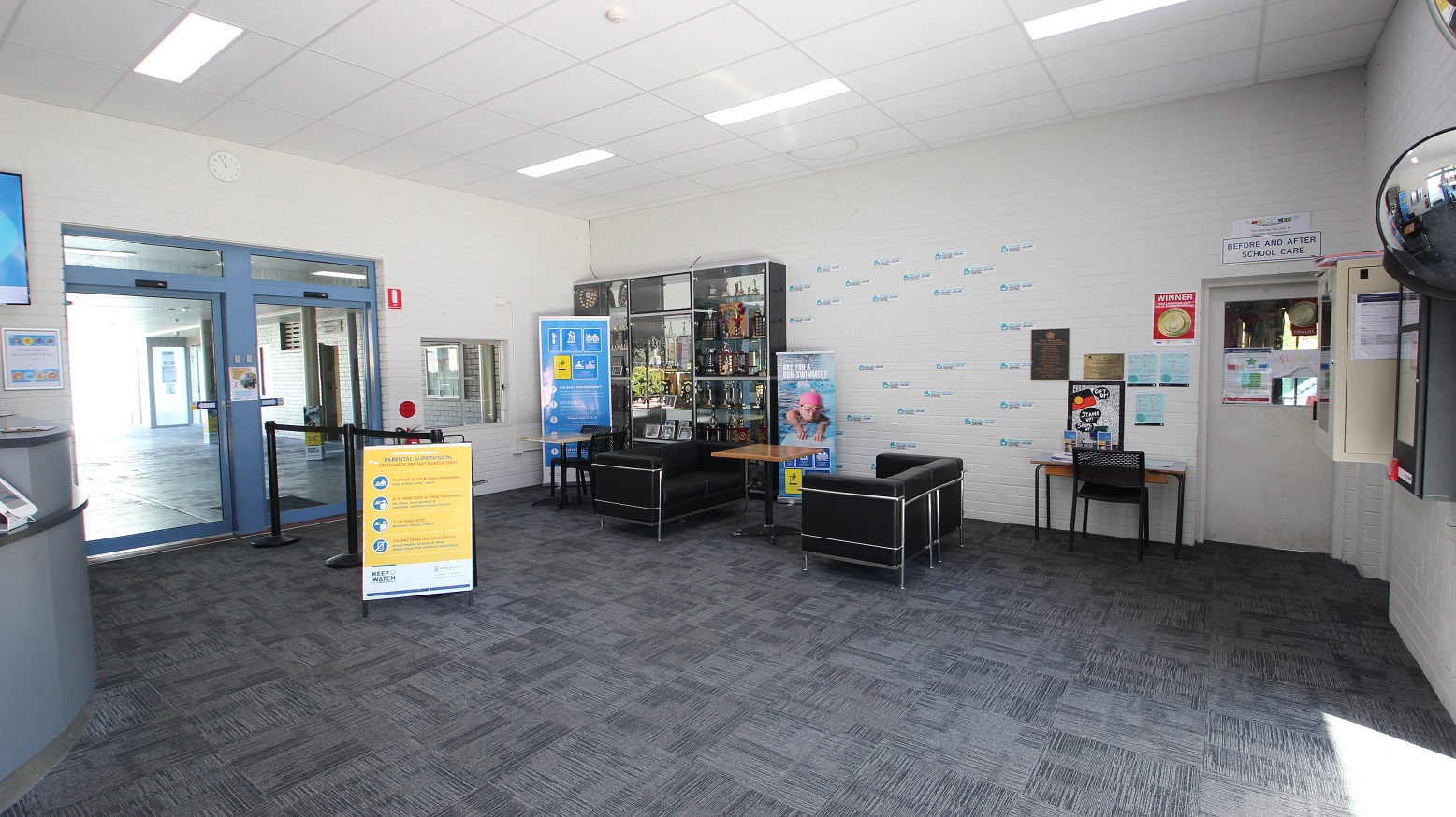 Opposite the curvy reception desk at the Michael Wendon Aquatic Leisure Centre is a seating area that contains black sofa chairs with a brown table in the middle. A grey cabinet storing lots of trophies is covered with glass. Several tickers can also be seen on the white-painted brick wall. On the right is a white door for “before and after school care” while on the left is a black stanchion with black ropes leading to a glass sliding double door with blue painted edges. Opposite the curvy reception desk at the Michael Wendon Aquatic Leisure Centre is a seating area that contains black sofa chairs with a brown table in the middle. A grey cabinet storing lots of trophies is covered with glass. Several tickers can also be seen on the white-painted brick wall. On the right is a white door for “before and after school care” while on the left is a black stanchion with black ropes leading to a glass sliding double door with blue painted edges.