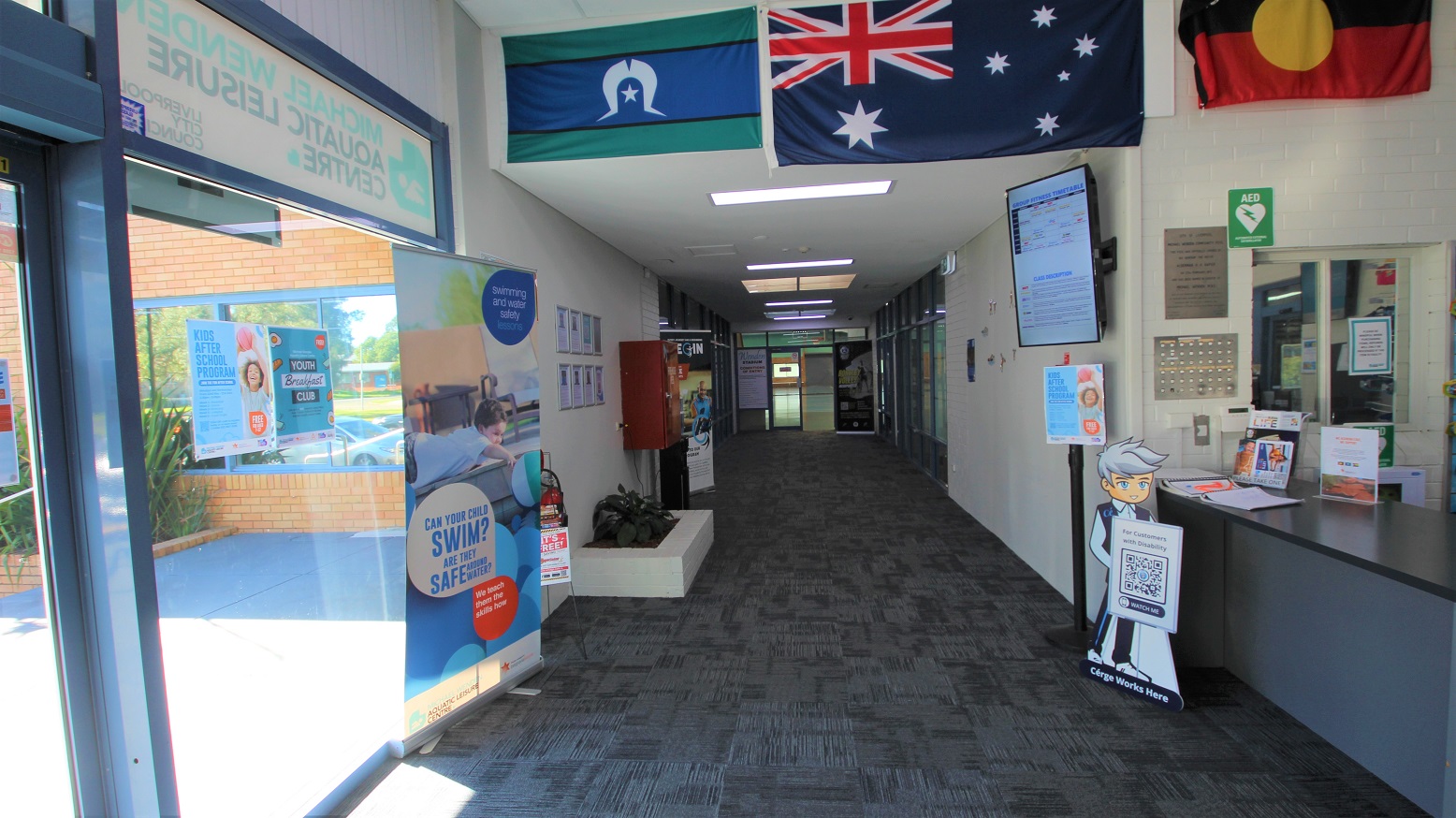 Hallway leading to the Stadium and Gymnasium areas at the Michael Wendon Aquatic Leisure Centre. In front of the reception desk is the brand icon of Cérge with a QR code to help customers with disabilities. The space before the ceiling is occupied by the Australian, Torres Strait, and Indigenous Australian flags. The television mounted on the right wall is in a vertical manner. On the left of this hallway, you can find a fire extinguisher and a small white plant box. Hallway leading to the Stadium and Gymnasium areas at the Michael Wendon Aquatic Leisure Centre. In front of the reception desk is the brand icon of Cérge with a QR code to help customers with disabilities. The space before the ceiling is occupied by the Australian, Torres Strait, and Indigenous Australian flags. The television mounted on the right wall is in a vertical manner. On the left of this hallway, you can find a fire extinguisher and a small white plant box.