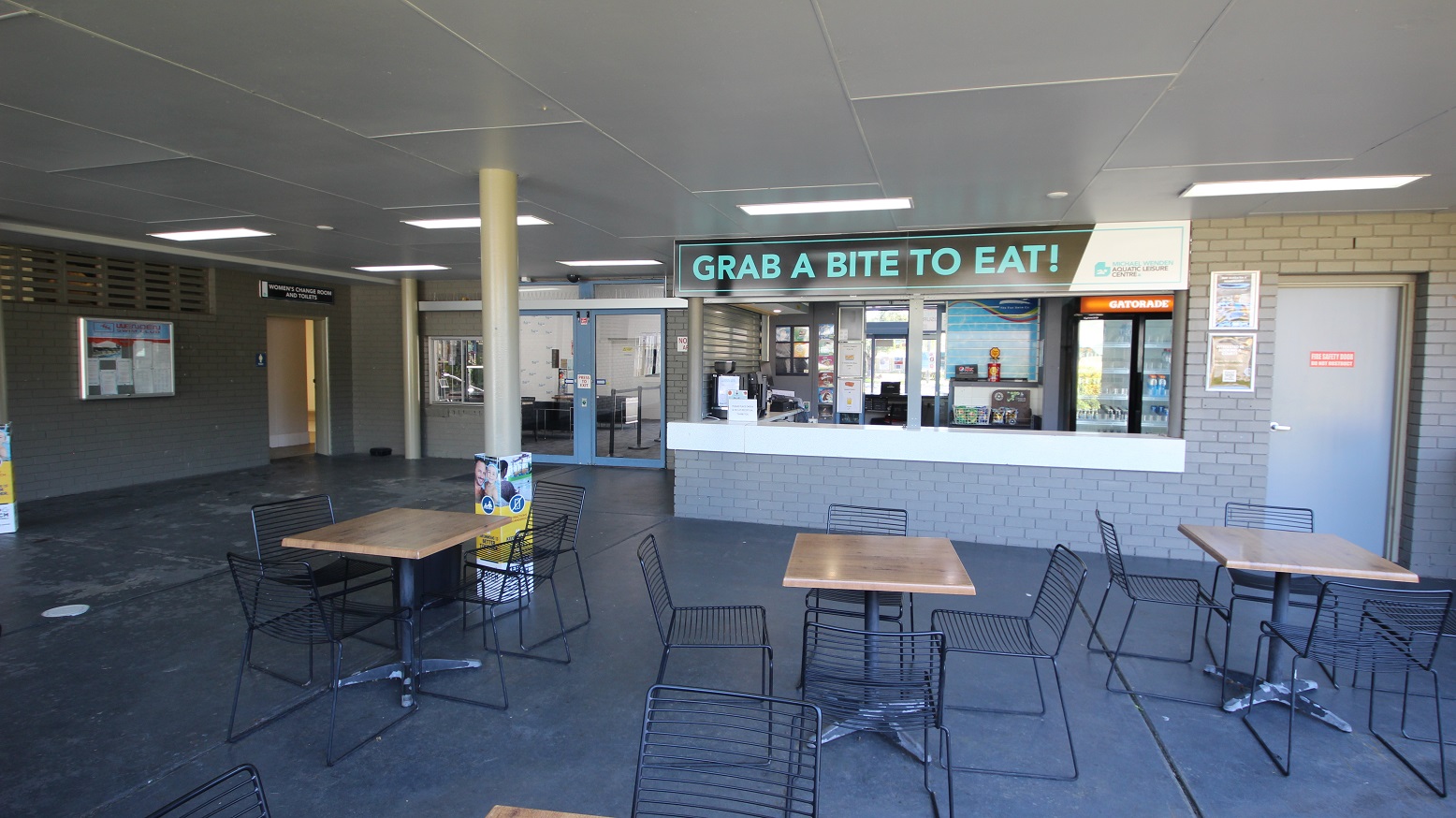 Café inside the Michael Wendon Aquatic Leisure Centre. Noticeable in the Café is the signboard which contains a neon blue, “GRAB A BITE TO EAT!”. The brick walls and floors are grey. On the left side is the entry point to the women’s change room and toilet beside a bulletin board. The seating area in the Café is composed of black metal frame chairs and wooden tables with grey support. Café inside the Michael Wendon Aquatic Leisure Centre. Noticeable in the Café is the signboard which contains a neon blue, “GRAB A BITE TO EAT!”. The brick walls and floors are grey. On the left side is the entry point to the women’s change room and toilet beside a bulletin board. The seating area in the Café is composed of black metal frame chairs and wooden tables with grey support.