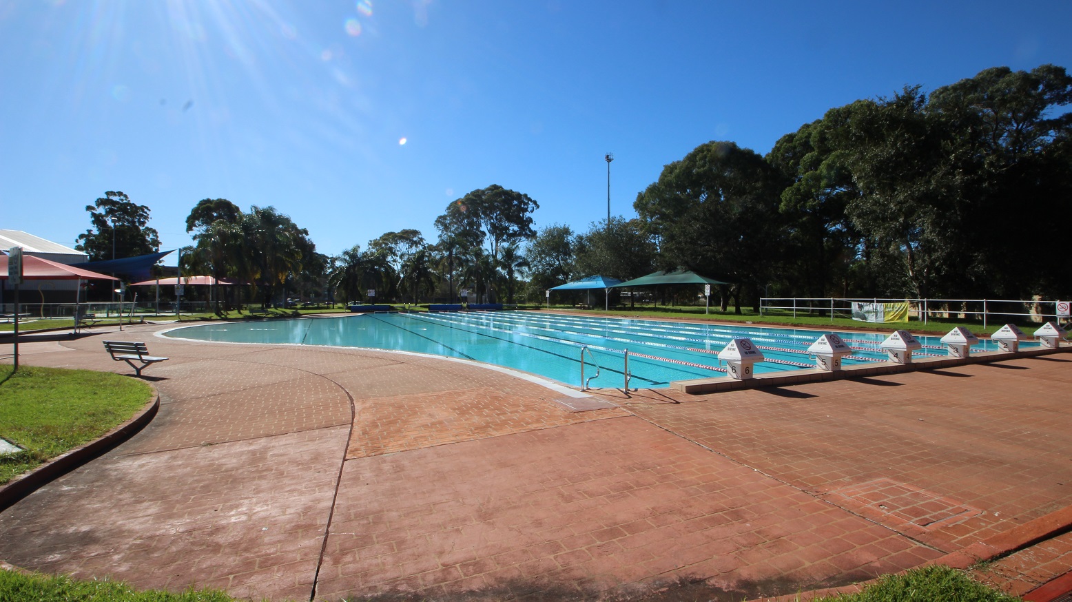 A closer view of the 50 meters outdoor pool located in the park-like outdoor area at Michael Wendon Aquatic Leisure Centre. The lanes are separated by white, blue, and red floating ropes. Surrounding the pool is a brick pathway to move around the area. On the left is a pool ladder. At the top of the slightly elevated section of the pool starting point is a mini-house-looking shape with a no-diving sign and a number etched on the bottom that increments from right to left. A closer view of the 50 meters outdoor pool located in the park-like outdoor area at Michael Wendon Aquatic Leisure Centre. The lanes are separated by white, blue, and red floating ropes. Surrounding the pool is a brick pathway to move around the area. On the left is a pool ladder. At the top of the slightly elevated section of the pool starting point is a mini-house-looking shape with a no-diving sign and a number etched on the bottom that increments from right to left.