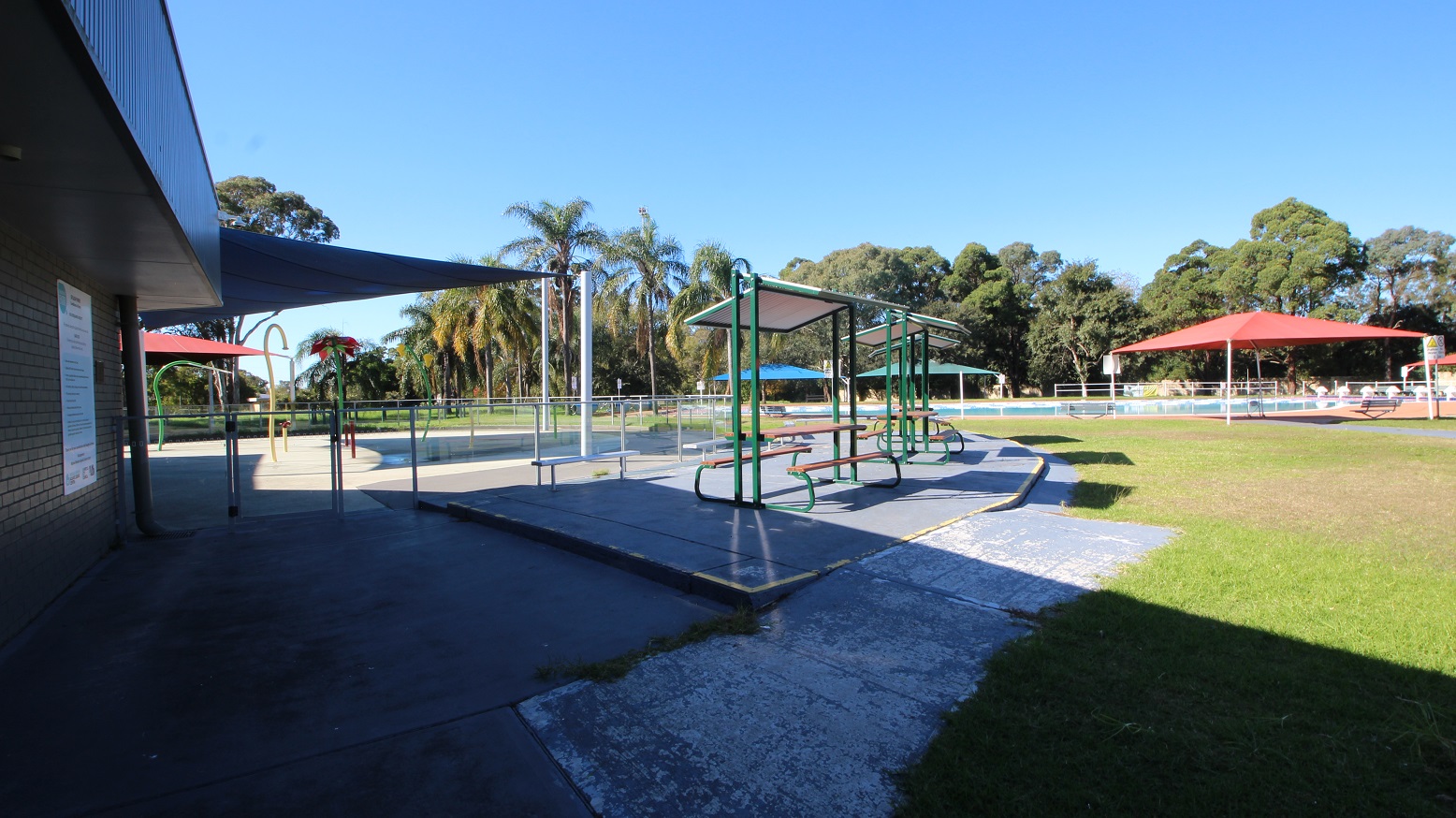 A closer look at the outdoor shelters near the 50 meters outdoor pool. It has a wooden table and seating, white roofing, and a green-painted metal frame. Behind the outdoor shelters is a hip-high glass fence with metal handrails enclosing a space that has yellow and green curvy tubes and bollards. A closer look at the outdoor shelters near the 50 meters outdoor pool. It has a wooden table and seating, white roofing, and a green-painted metal frame. Behind the outdoor shelters is a hip-high glass fence with metal handrails enclosing a space that has yellow and green curvy tubes and bollards.