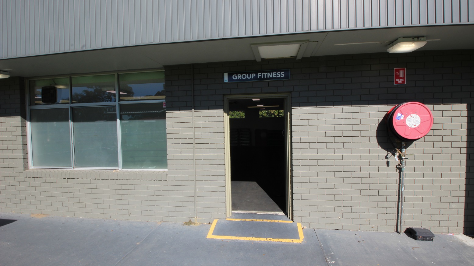 The entrance door to the Group Fitness room at Michael Wendon Aquatic Leisure Centre can be identified by a yellow rectangular marking drawn on the floor. On the right wall is a red fire hose mounted on the grey-painted brick wall. There is a glass window on the left. The entrance door to the Group Fitness room at Michael Wendon Aquatic Leisure Centre can be identified by a yellow rectangular marking drawn on the floor. On the right wall is a red fire hose mounted on the grey-painted brick wall. There is a glass window on the left.