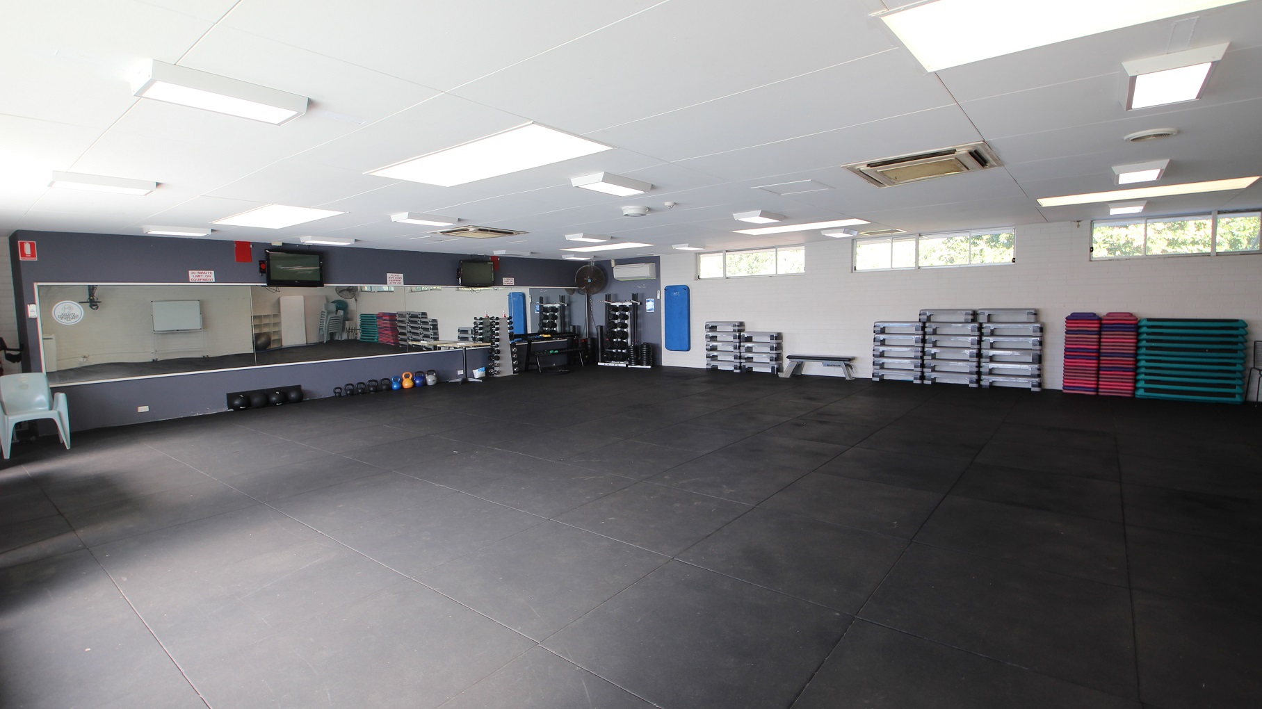 The Group Fitness room has a large open space with a grey rubber mat. The front wall with a medium size mirror is painted grey while the other walls are painted white. On the floor underneath the mirror are body bars in different colors while on the left side is a large white chair. On the upper part of the white-painted wall are small, sliding glass windows and a set of grey platforms stacked on top of each other. The Group Fitness room has a large open space with a grey rubber mat. The front wall with a medium size mirror is painted grey while the other walls are painted white. On the floor underneath the mirror are body bars in different colors while on the left side is a large white chair. On the upper part of the white-painted wall are small, sliding glass windows and a set of grey platforms stacked on top of each other.