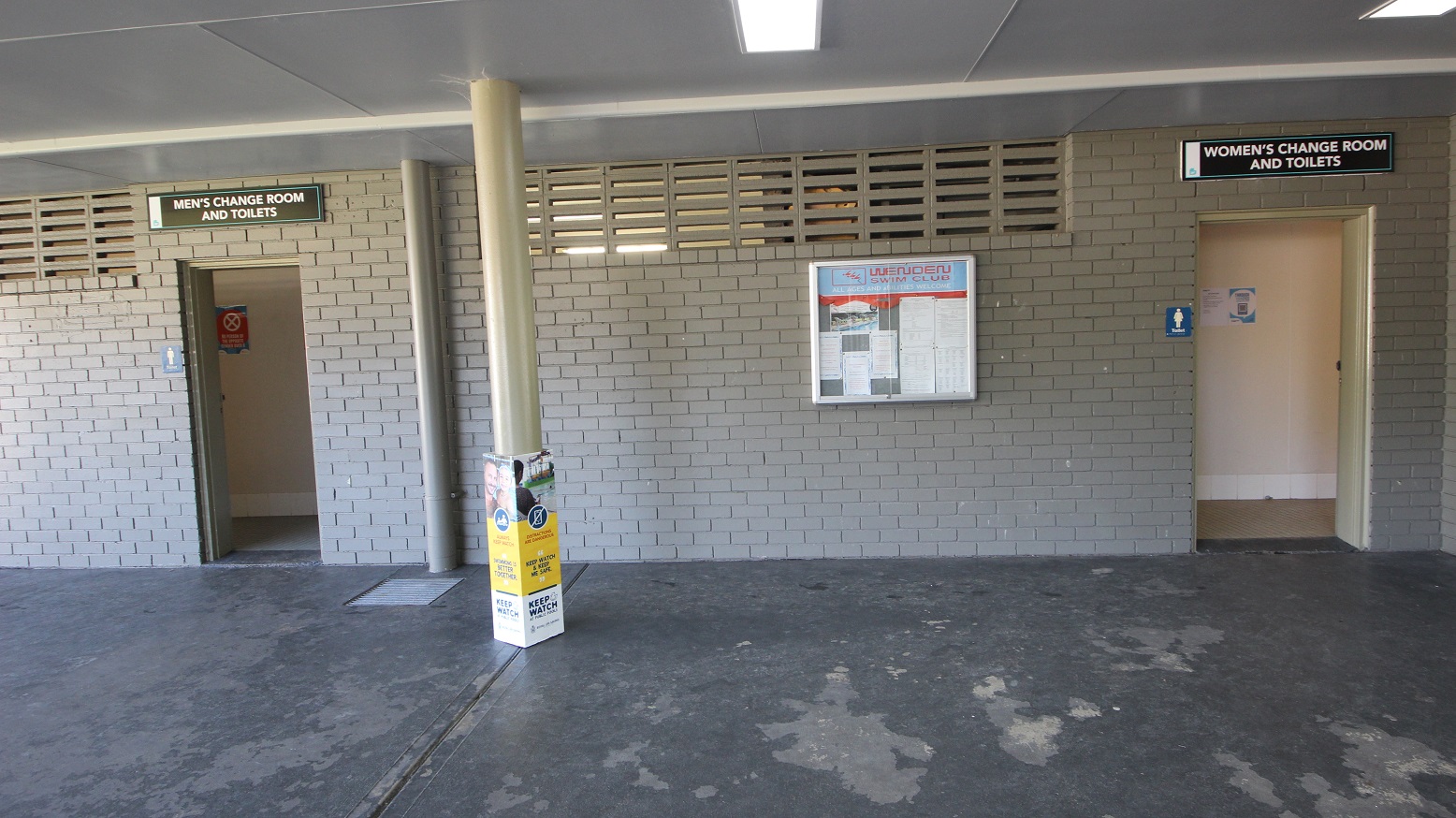 The image shows the men’s change room and toilets on the left side while the women’s change room and toilets are on the right. The brick wall is painted grey and on some parts, on the upper side of the wall, is a column of narrow rectangular holes. Pinned inside the bulletin board located between the wall of the toilet doors are white papers with fine prints. The image shows the men’s change room and toilets on the left side while the women’s change room and toilets are on the right. The brick wall is painted grey and on some parts, on the upper side of the wall, is a column of narrow rectangular holes. Pinned inside the bulletin board located between the wall of the toilet doors are white papers with fine prints.