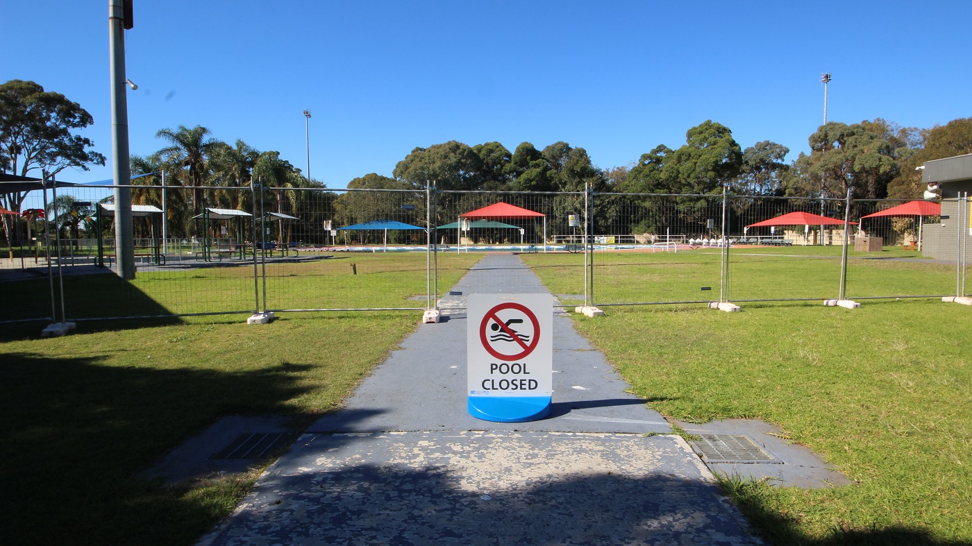 Pool closed sign board placed on a grey, smooth concrete pathway leading to the 50 meters outdoor pool. The area is surrounded by a tall metal fence with small square holes, which gives guests a view of the surrounding area. The large tents, outdoor shelters, palm, and other tree types are visible in this area. Pool closed sign board placed on a grey, smooth concrete pathway leading to the 50 meters outdoor pool. The area is surrounded by a tall metal fence with small square holes, which gives guests a view of the surrounding area. The large tents, outdoor shelters, palm, and other tree types are visible in this area.