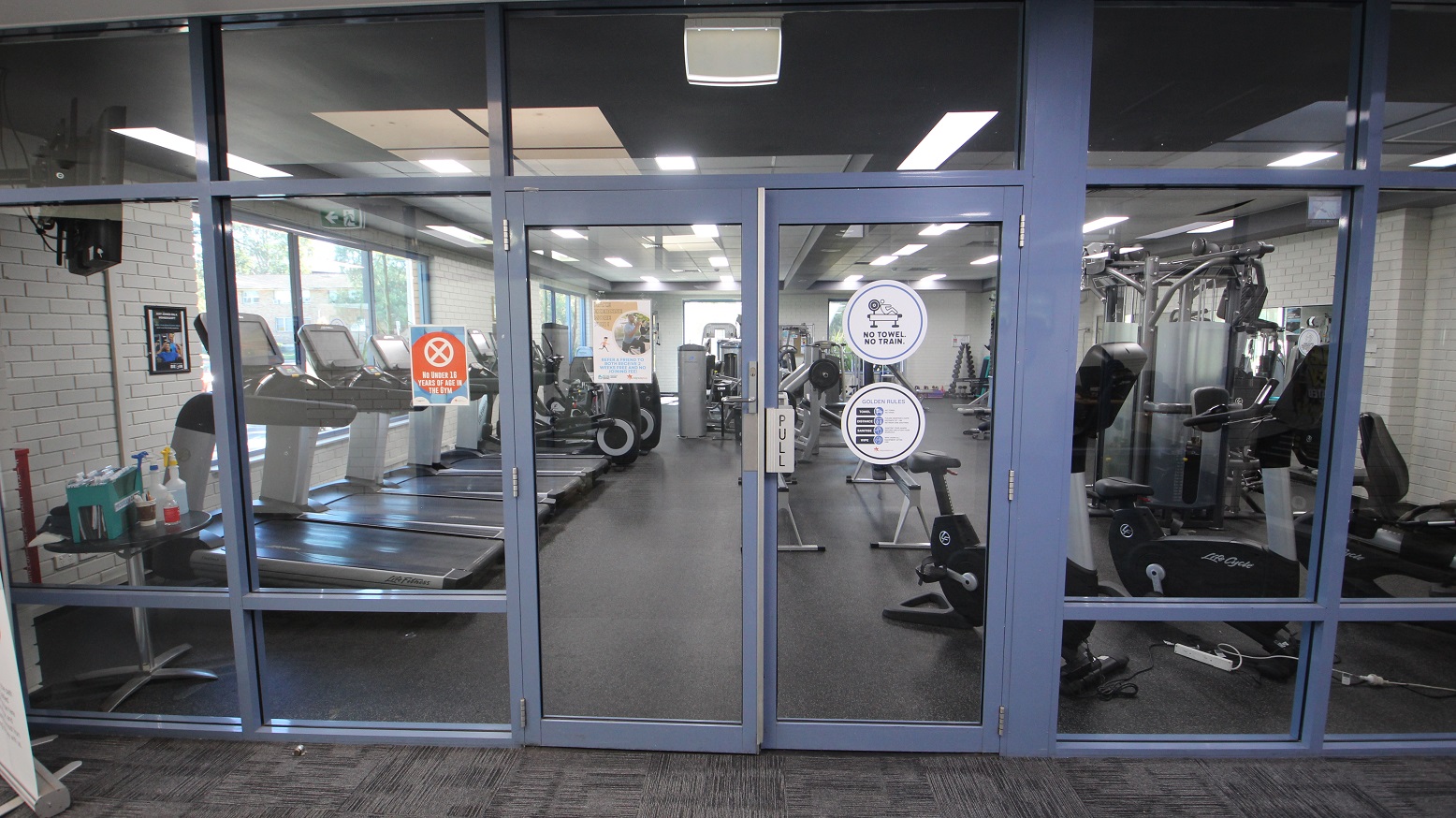 Front of the manually operated glass doors with blue painted edges of the Gymnasium at the Michael Wendon Aquatic Leisure Centre. Opening the door is by pulling it towards yourself. A warning sign on the orange poster on the left prohibits those under 16 years of age from entering this area. Rules and conditions before entering the gymnasium can be found on the right of the entrance door. Seen through the glass doors are cardio and weight training equipment. Front of the manually operated glass doors with blue painted edges of the Gymnasium at the Michael Wendon Aquatic Leisure Centre. Opening the door is by pulling it towards yourself. A warning sign on the orange poster on the left prohibits those under 16 years of age from entering this area. Rules and conditions before entering the gymnasium can be found on the right of the entrance door. Seen through the glass doors are cardio and weight training equipment.
