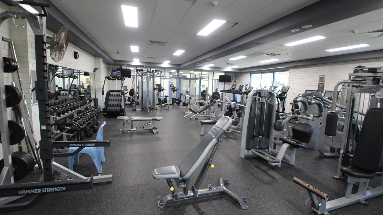 Opposite view inside the gymnasium. This image shows a closer look of the weight training equipment available inside the Gymnasium at Michael Wendon Aquatic Leisure Centre. There is a mirror behind the dumbbell rack and on the upper part is a black analog clock. In front of the wall with a black fan is a large blue plastic chair. Opposite view inside the gymnasium. This image shows a closer look of the weight training equipment available inside the Gymnasium at Michael Wendon Aquatic Leisure Centre. There is a mirror behind the dumbbell rack and on the upper part is a black analog clock. In front of the wall with a black fan is a large blue plastic chair.