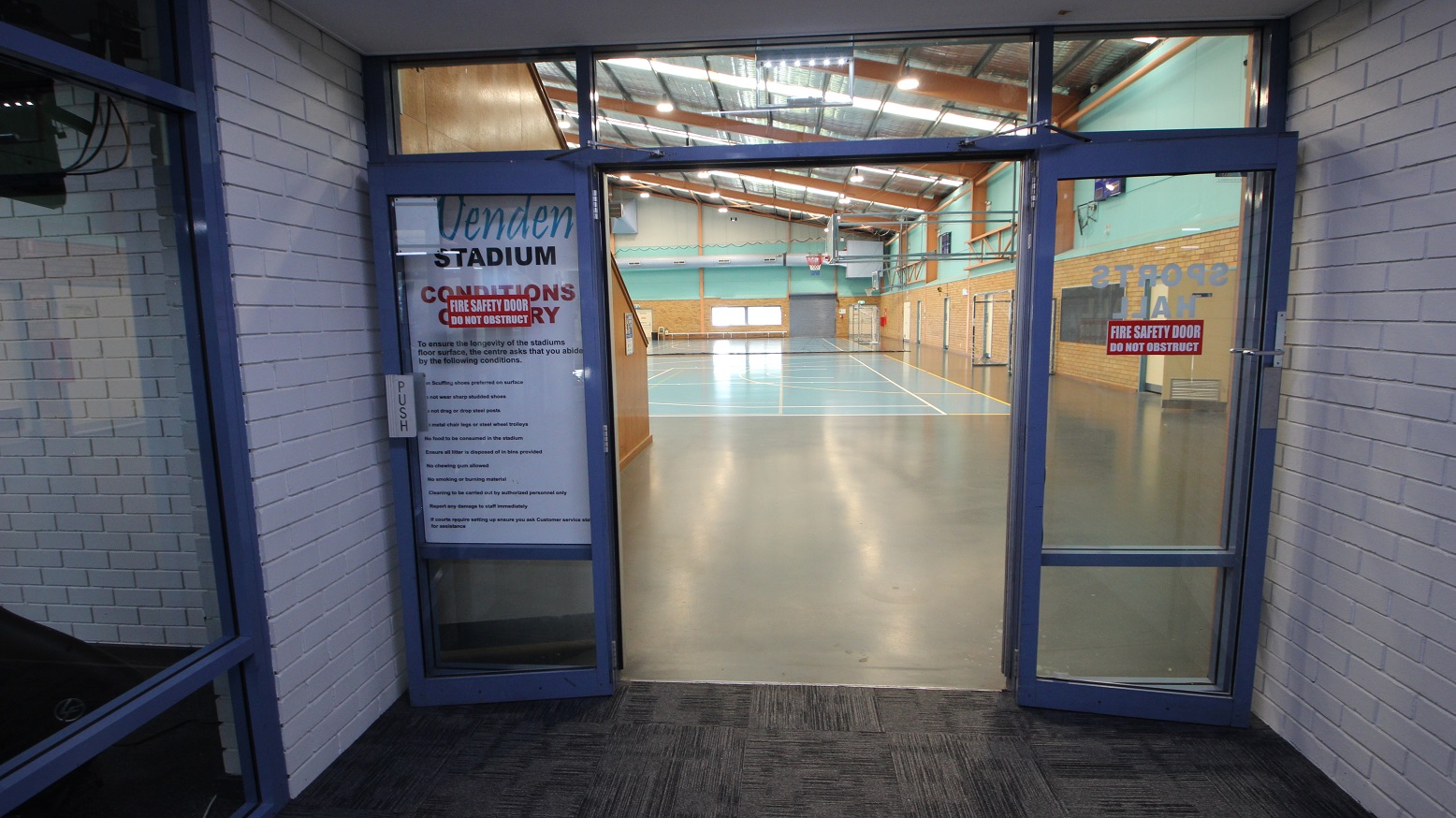 Open doors of the Stadium at the Michael Wendon Aquatic Leisure Centre. The Stadium has a manually operated, glass double door with blue painted edges. The right portion of the Stadium seen through the entry doors shows a sky-blue concrete floor with white & yellow lines. The lower section of the wall is made up of brown-painted bricks, while the upper section is painted sky blue. The Stadium has an insulation-lined roof with skylights and apricot-colored beams. Open doors of the Stadium at the Michael Wendon Aquatic Leisure Centre. The Stadium has a manually operated, glass double door with blue painted edges. The right portion of the Stadium seen through the entry doors shows a sky-blue concrete floor with white & yellow lines. The lower section of the wall is made up of brown-painted bricks, while the upper section is painted sky blue. The Stadium has an insulation-lined roof with skylights and apricot-colored beams.