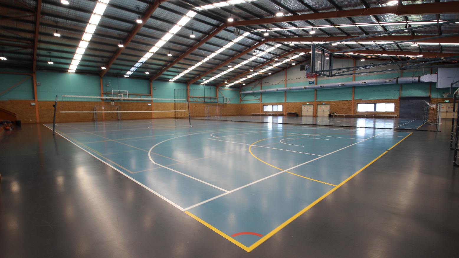 Entire view of the Stadium at the Michael Wendon Aquatic Leisure Centre. The sporting stadium is made of two basketball courts in size and is separated by a black net in the middle. The first court near the entrance doors has a volleyball net while on the second court is a white football net. Scattered near the brick walls and glass windows are long benches. There are dirty-white double doors with horizontal metal handles at one end of this stadium. Entire view of the Stadium at the Michael Wendon Aquatic Leisure Centre. The sporting stadium is made of two basketball courts in size and is separated by a black net in the middle. The first court near the entrance doors has a volleyball net while on the second court is a white football net. Scattered near the brick walls and glass windows are long benches. There are dirty-white double doors with horizontal metal handles at one end of this stadium.