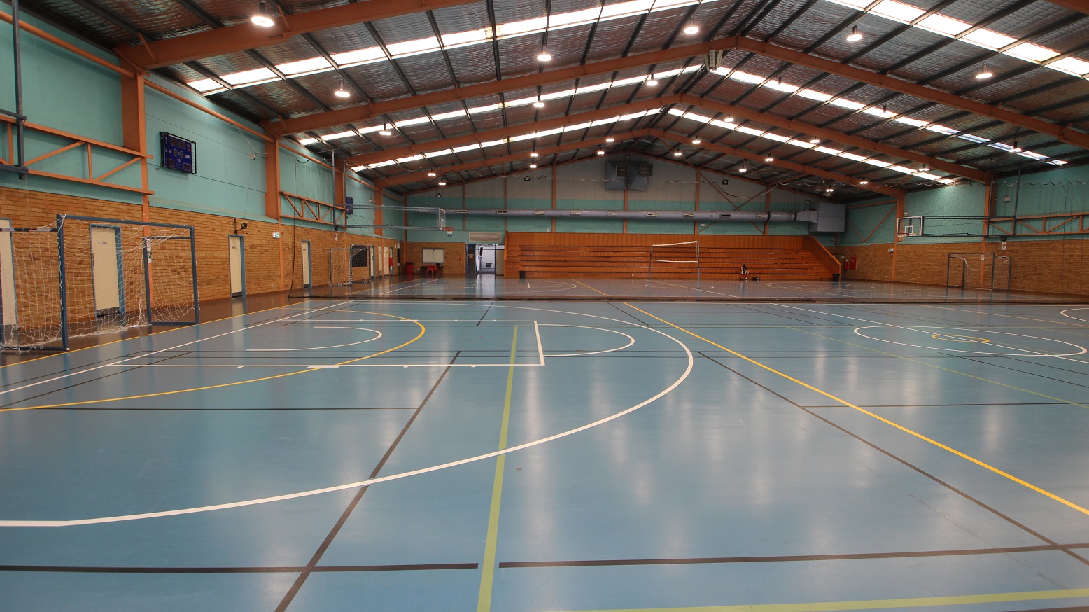 A closer look at the second court of the Stadium at the Michael Wendon Aquatic Leisure Centre. The white football net is wrapped in a grey metal frame. Aside from the white and yellow lines, there is also a black line visible on the floor of this court. There is a six-tiered stadium seating made out of timber situated to the left of the entrance doors. A closer look at the second court of the Stadium at the Michael Wendon Aquatic Leisure Centre. The white football net is wrapped in a grey metal frame. Aside from the white and yellow lines, there is also a black line visible on the floor of this court. There is a six-tiered stadium seating made out of timber situated to the left of the entrance doors.
