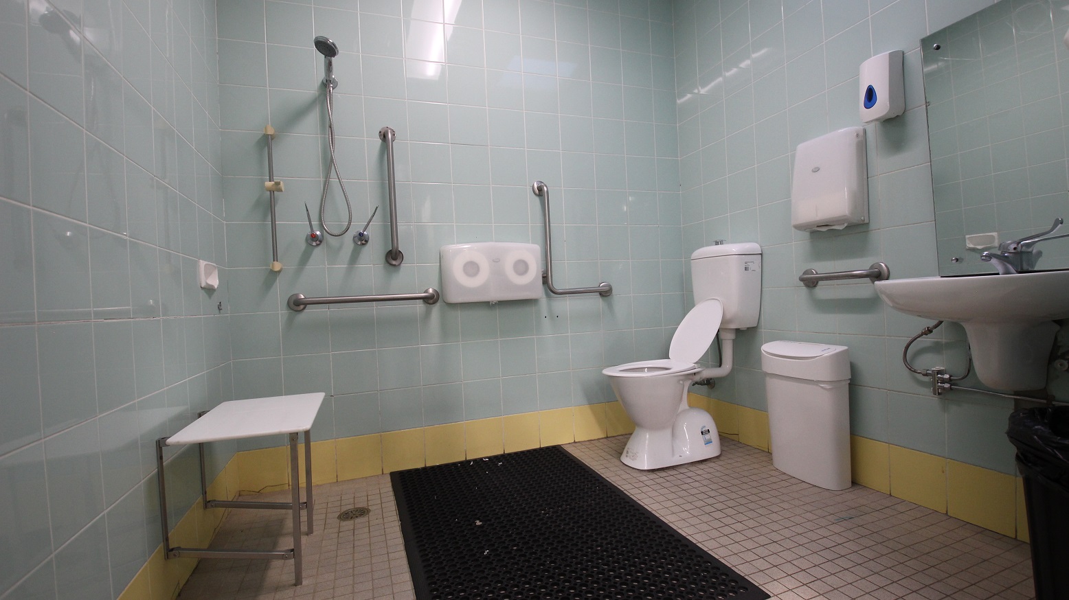 View inside the Accessible bathroom in the Gymnasium at the Michael Wendon Aquatic Leisure Centre. It has a black rubber mat, a wash basin with a mirror, an automatic liquid dispenser mounted on the light blue tiled wall, a white and black rubbish bin, two rolls of tissue papers, a handheld shower, and grab bars. The tiles at the lowermost part of the wall are painted in yellow. View inside the Accessible bathroom in the Gymnasium at the Michael Wendon Aquatic Leisure Centre. It has a black rubber mat, a wash basin with a mirror, an automatic liquid dispenser mounted on the light blue tiled wall, a white and black rubbish bin, two rolls of tissue papers, a handheld shower, and grab bars. The tiles at the lowermost part of the wall are painted in yellow.