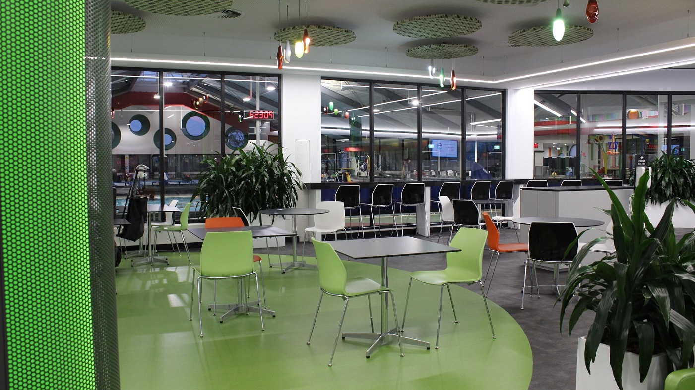 Apart from the black and white tables and chairs in the seating area in front of the Cafe are the green and orange chairs with steel legs. Potted plants are strategically placed around the seating area. Colorful bulbs with tear shapes and circular flat baskets with black and green stripes can be seen on the ceiling. The pool hall is visible through the glass walls.