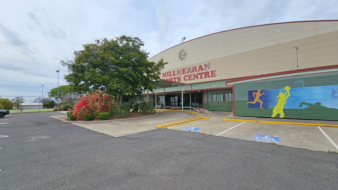 Millmerran Sports Centre with a large sign on the building. On the right, a mural depicts colourful silhouettes of athletes. The entrance is in the centre, with a covered walkway. On the left, there are trees and shrubs, and a car is parked in the distance. The foreground shows a paved area with a marked disabled parking space.