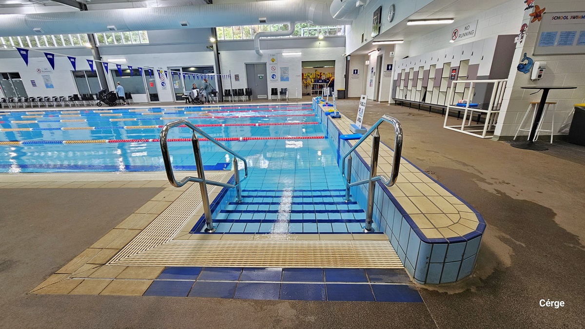 Pool stair with handrails of the 25m, 6-lane heated indoor pool. Each lane is separated by white, yellow, red, and blue floating rope lanes. At the right pool coping are white sign boards with small blue rectangular blocks to hold its position. Overhang perpendicular to the pool is a string of triangular blue flags. Against the right white wall is a white and grey cabinet locker. Above it, there is a large white sign for NO RUNNING. To the back is an open space filled with yellow life jackets. Pool stair with handrails of the 25m, 6-lane heated indoor pool. Each lane is separated by white, yellow, red, and blue floating rope lanes. At the right pool coping are white sign boards with small blue rectangular blocks to hold its position. Overhang perpendicular to the pool is a string of triangular blue flags. Against the right white wall is a white and grey cabinet locker. Above it, there is a large white sign for NO RUNNING. To the back is an open space filled with yellow life jackets.