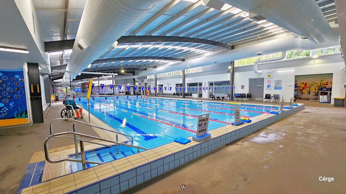 25-metre Heated Indoor Pool at Monbulk Aquatic Centre. Near the white tile pool at the starting line, to the left, are stairs with metal handrails. The pool deck is concrete in color. The facility has white walls, large white ventilation ducts, and grey metal beams in the ceiling. On the upper right wall are small glass windows that allow natural light to filter in. Metal benches and black plastic chairs are available around the pool deck. There is an aquatic chair on the left pool deck. 25-metre Heated Indoor Pool at Monbulk Aquatic Centre. Near the white tile pool at the starting line, to the left, are stairs with metal handrails. The pool deck is concrete in color. The facility has white walls, large white ventilation ducts, and grey metal beams in the ceiling. On the upper right wall are small glass windows that allow natural light to filter in. Metal benches and black plastic chairs are available around the pool deck. There is an aquatic chair on the left pool deck.