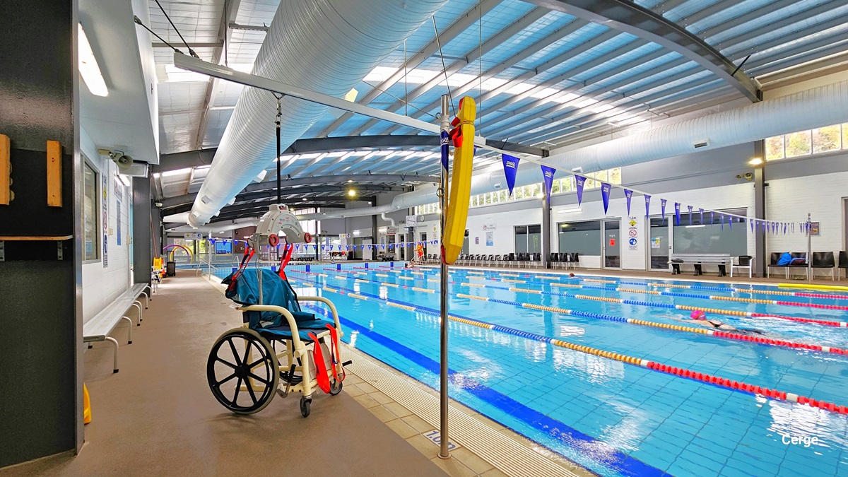 Left pool deck of 25m, 6-lane heated indoor pool with an aquatic wheelchair that has blue seating and red straps supported by a grey hoist with black rope. Found against the left white brick wall is a long, grey metal bench. On the pool coping, within the floor with a metal pole that has a yellow floating device, is a white 1.0m sign for water depth. On the bottom pool floor is the bold blue center T line that helps the swimmer stay at the center. On the walls are different precautionary signs. Left pool deck of 25m, 6-lane heated indoor pool with an aquatic wheelchair that has blue seating and red straps supported by a grey hoist with black rope. Found against the left white brick wall is a long, grey metal bench. On the pool coping, within the floor with a metal pole that has a yellow floating device, is a white 1.0m sign for water depth. On the bottom pool floor is the bold blue center T line that helps the swimmer stay at the center. On the walls are different precautionary signs.