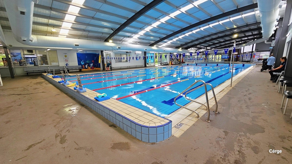 On the right pool deck of 25m, 6-lane heated indoor pool, are two stainless steel grab rails. The right pool coping has a small white sign with a blue 0.9m sign for water depth. The elevated pool coping at the start is white sign boards with small blue rectangular blocks to hold its position. Behind the long grey metal bench of the opposite pool deck is a glass window that shows a part of the café, warning signs for pool rules, and a blue mural with different sea creatures. On the right pool deck of 25m, 6-lane heated indoor pool, are two stainless steel grab rails. The right pool coping has a small white sign with a blue 0.9m sign for water depth. The elevated pool coping at the start is white sign boards with small blue rectangular blocks to hold its position. Behind the long grey metal bench of the opposite pool deck is a glass window that shows a part of the café, warning signs for pool rules, and a blue mural with different sea creatures.