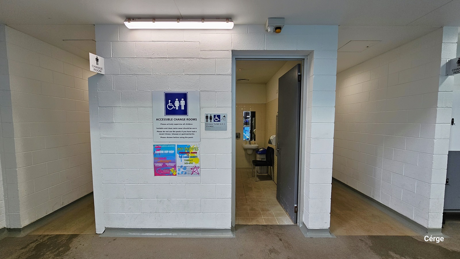 Accessible Change Room sign placed on a white brick wall at Monbulk Aquatic Center. A fluorescent ceiling illuminates the front of the change room. Underneath the Accessible Change Rooms sign are two colorful advertisements. On the right is a grey metal plate for the Unisex Toilet LH + Shower sign. Seen through the open grey metal door is the black plastic chair, wash basin with mirror, and a grey garbage bin with a blue lid. It has a beige tile wall and a brown tile floor. Accessible Change Room sign placed on a white brick wall at Monbulk Aquatic Center. A fluorescent ceiling illuminates the front of the change room. Underneath the Accessible Change Rooms sign are two colorful advertisements. On the right is a grey metal plate for the Unisex Toilet LH + Shower sign. Seen through the open grey metal door is the black plastic chair, wash basin with mirror, and a grey garbage bin with a blue lid. It has a beige tile wall and a brown tile floor.