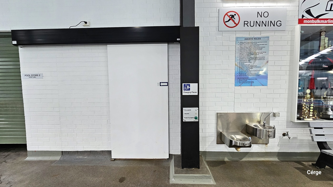 Changing Places at Monbulk Aquatic Centre has a white sliding door with an arrow pointing to the left placed within a white brick wall with black wall lining. The door can be operated through a grey metal plate that contains the green button for opening the door and two lights for occupied and vacant. On the right wall is a stainless steel, wall-mounted washbasin and drinking fountain and above it is a white NO RUNNING sign and blue aquatic rules. To the right is a glass display of trophies. Changing Places at Monbulk Aquatic Centre has a white sliding door with an arrow pointing to the left placed within a white brick wall with black wall lining. The door can be operated through a grey metal plate that contains the green button for opening the door and two lights for occupied and vacant. On the right wall is a stainless steel, wall-mounted washbasin and drinking fountain and above it is a white NO RUNNING sign and blue aquatic rules. To the right is a glass display of trophies.