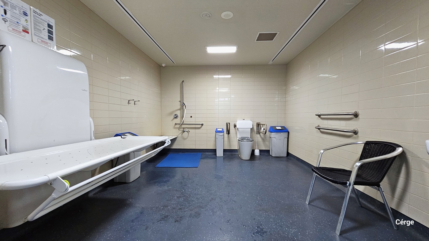 Inside the changing place is a white changing table attached to a rectangular white platform on the left. Next to it is a grey garbage bin with a blue lid, a mounted white shower seat, and a shower section with a handheld shower head and blue rubber mat. The toilet has a grey seat cover and two curved metal grab bars, as well as grey garbage bins with blue lids. On the right wall is a black plastic chair and two grab bars. The room has a dark grey floor, beige tile walls, and a white ceiling. Inside the changing place is a white changing table attached to a rectangular white platform on the left. Next to it is a grey garbage bin with a blue lid, a mounted white shower seat, and a shower section with a handheld shower head and blue rubber mat. The toilet has a grey seat cover and two curved metal grab bars, as well as grey garbage bins with blue lids. On the right wall is a black plastic chair and two grab bars. The room has a dark grey floor, beige tile walls, and a white ceiling.