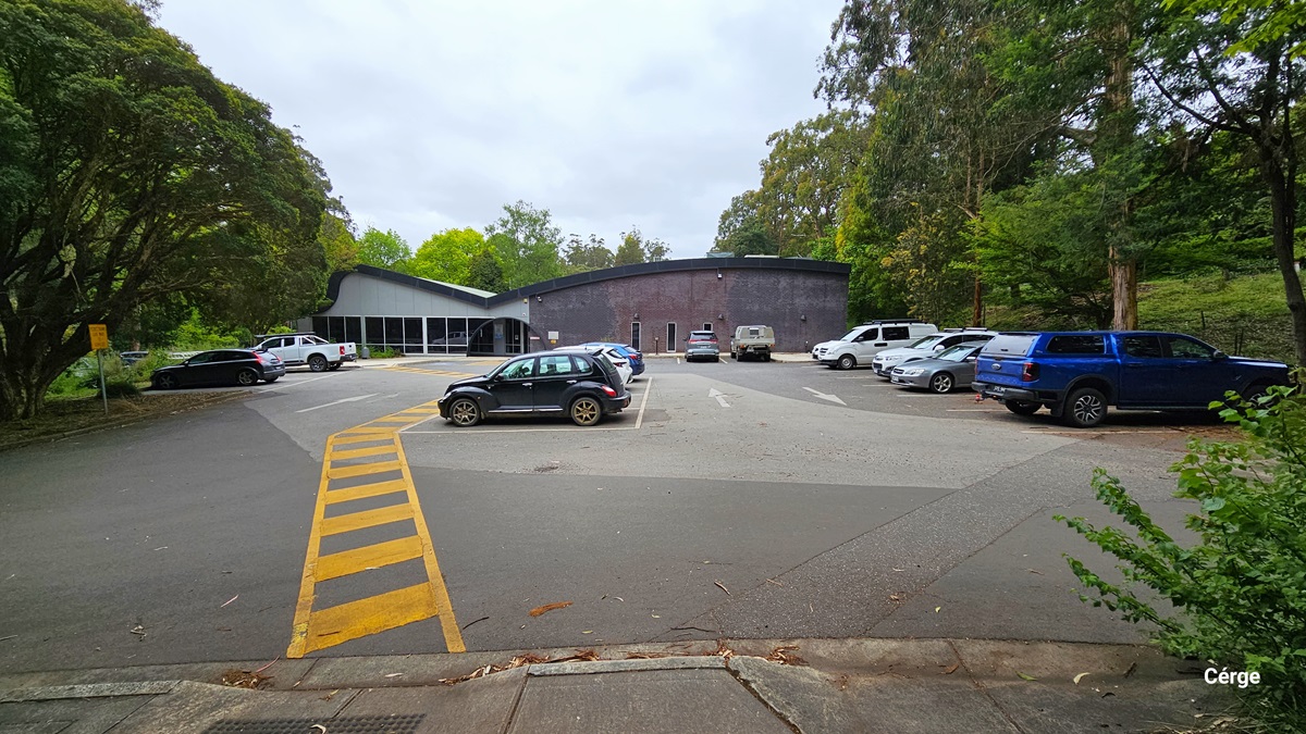 Parking spaces in Monbulk Aquatic Centre nestled in a wooden area. The park space floor transitions from dark grey to light grey and back to dark grey when going closer to the entrance. On the floor surface of the parking area are white directional arrows and yellow lines marking the parking spaces. Monbulk Aquatic Center has a curved roof with grey to dark purple brick wall finish. The entrance can be distinguished by a black arch near the glass windows with white metal frames. Parking spaces in Monbulk Aquatic Centre nestled in a wooden area. The park space floor transitions from dark grey to light grey and back to dark grey when going closer to the entrance. On the floor surface of the parking area are white directional arrows and yellow lines marking the parking spaces. Monbulk Aquatic Center has a curved roof with grey to dark purple brick wall finish. The entrance can be distinguished by a black arch near the glass windows with white metal frames.