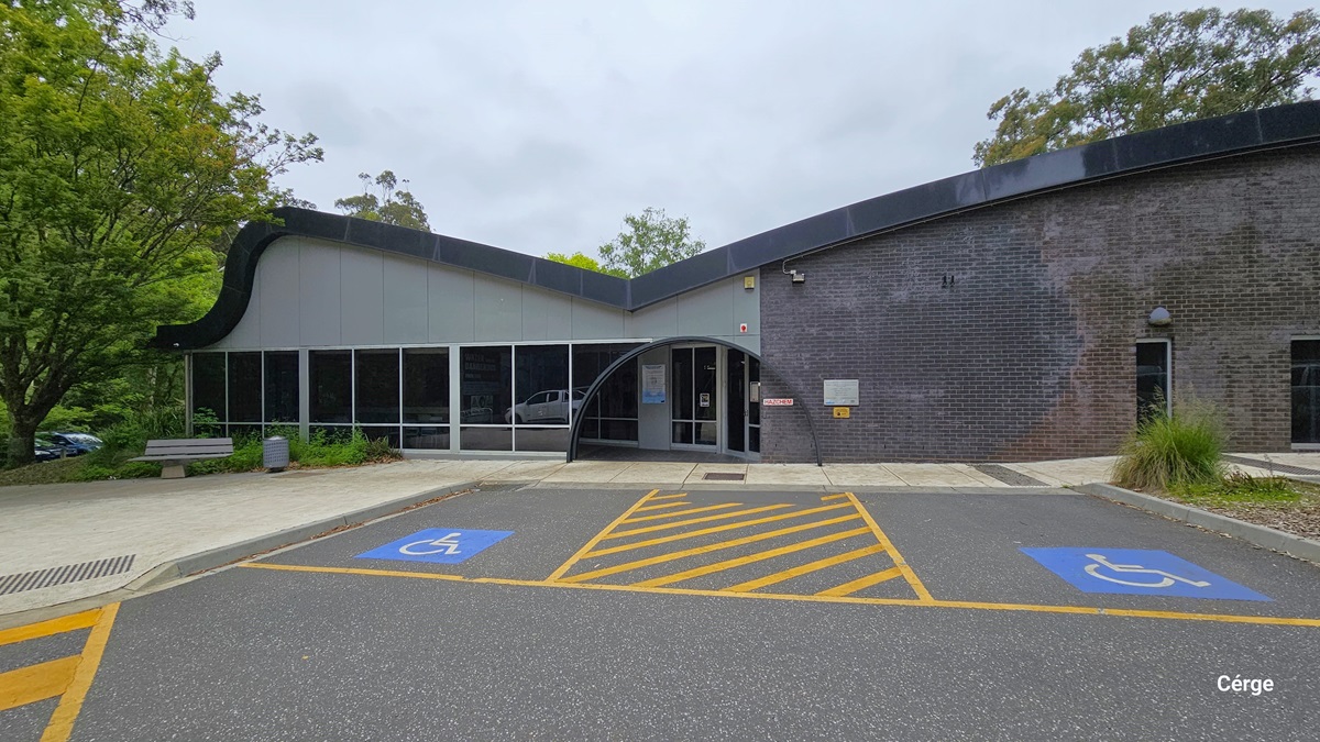 Two accessible car parking spaces in front of Monbulk Aquatic Centre are marked by a blue background with a white wheelchair image. Between the 2 accessible car park spaces are yellow diagonal lines enclosed by a yellow rectangle. The building has a large glass window on the left side and a smaller window on the right side. The entrance is in the center of the building and has a black arch above it. Two accessible car parking spaces in front of Monbulk Aquatic Centre are marked by a blue background with a white wheelchair image. Between the 2 accessible car park spaces are yellow diagonal lines enclosed by a yellow rectangle. The building has a large glass window on the left side and a smaller window on the right side. The entrance is in the center of the building and has a black arch above it.