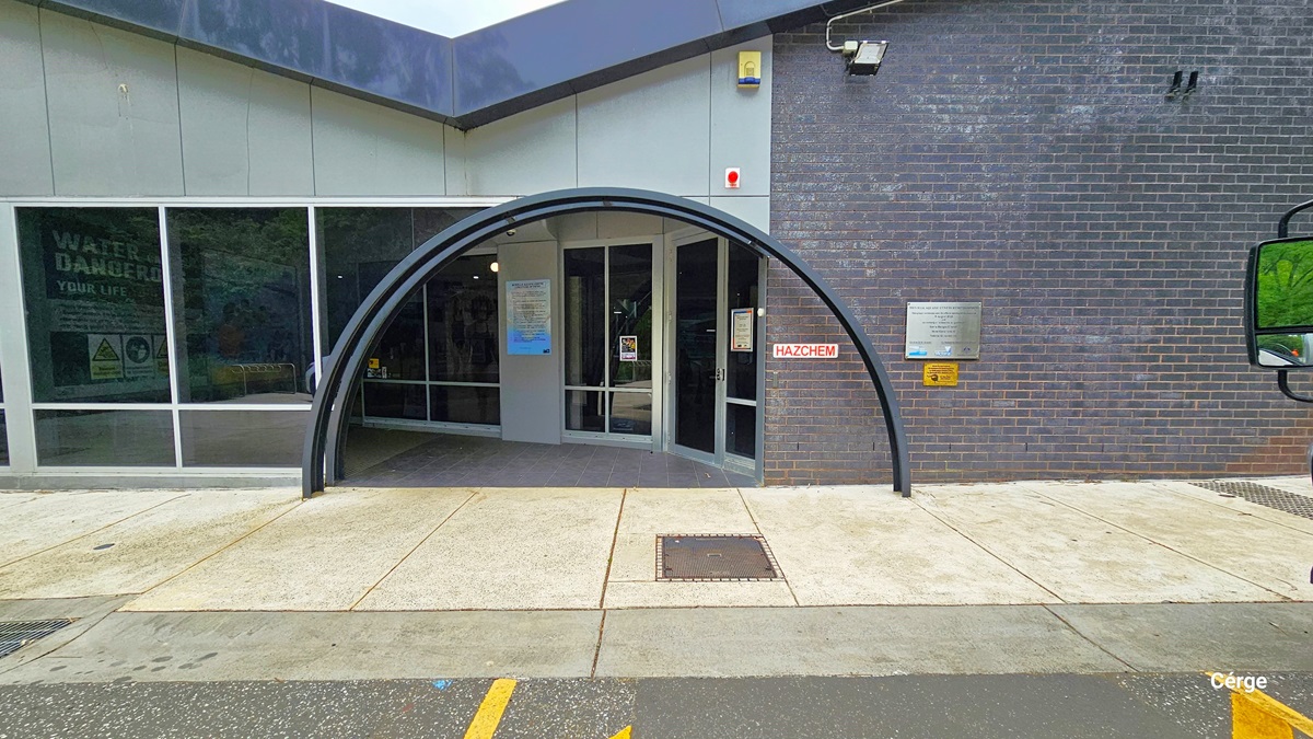 Entrance with two black metal arches at Monbulk Aquatic Centre. The entrance is a curved archway with a glass door and windows. The building transitions from grey tile with glass windows that have white metal frames to dark purple brick from left to right. The pathway in front is made of concrete tiles and is connected to the grey tiles of the entrance floor. A white sign on the brick wall has red HAZCHEM detailed on it. Entrance with two black metal arches at Monbulk Aquatic Centre. The entrance is a curved archway with a glass door and windows. The building transitions from grey tile with glass windows that have white metal frames to dark purple brick from left to right. The pathway in front is made of concrete tiles and is connected to the grey tiles of the entrance floor. A white sign on the brick wall has red HAZCHEM detailed on it.
