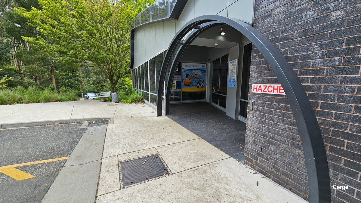 The side view of the entrance with two metal curved archways. The entrance is flanked by two large windows, one of which has a colorful mural on it. The entrance is also marked by a red and white “HAZCHEM” sign. The building is surrounded by lush greenery and a concrete walkway. Under the tall tree with a large tree canopy cover is a grey metal bench, and beside it is a grey circular perforated garbage bin. The side view of the entrance with two metal curved archways. The entrance is flanked by two large windows, one of which has a colorful mural on it. The entrance is also marked by a red and white “HAZCHEM” sign. The building is surrounded by lush greenery and a concrete walkway. Under the tall tree with a large tree canopy cover is a grey metal bench, and beside it is a grey circular perforated garbage bin.