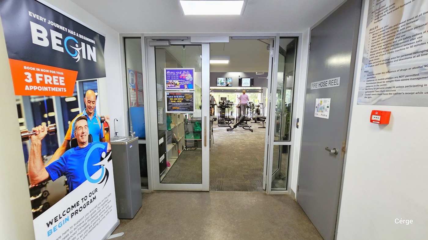 The gym entrance is through a set of double glass doors with white frames. The floor is of natural stone grey, while the walls and ceiling are white. On the ceiling just before the entrance door is a square fluorescent light. On the left side is a large poster detailing what the Begin Program is all about and a grey water fountain. On the left side is a grey door with a grey sign for FIRE HOSE REEL and a large board for facility rules. Seen through the open doors is a woman using an air walker. The gym entrance is through a set of double glass doors with white frames. The floor is of natural stone grey, while the walls and ceiling are white. On the ceiling just before the entrance door is a square fluorescent light. On the left side is a large poster detailing what the Begin Program is all about and a grey water fountain. On the left side is a grey door with a grey sign for FIRE HOSE REEL and a large board for facility rules. Seen through the open doors is a woman using an air walker.