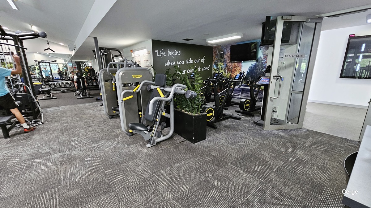 On the glass door that is open inward is a grey sign with black HEALTH CLUB letters. The door has a grey metal frame with a stainless steel U-shape handle. Behind the open glass door is a digital screen. The next section is for the black stationary bikes that have black walls with a motivational quote and a flat-screen TV mounted on the wall painted with a road and trees on the side. The other section has bench presses, dumbbells, and a lat pulldown. On the glass door that is open inward is a grey sign with black HEALTH CLUB letters. The door has a grey metal frame with a stainless steel U-shape handle. Behind the open glass door is a digital screen. The next section is for the black stationary bikes that have black walls with a motivational quote and a flat-screen TV mounted on the wall painted with a road and trees on the side. The other section has bench presses, dumbbells, and a lat pulldown.