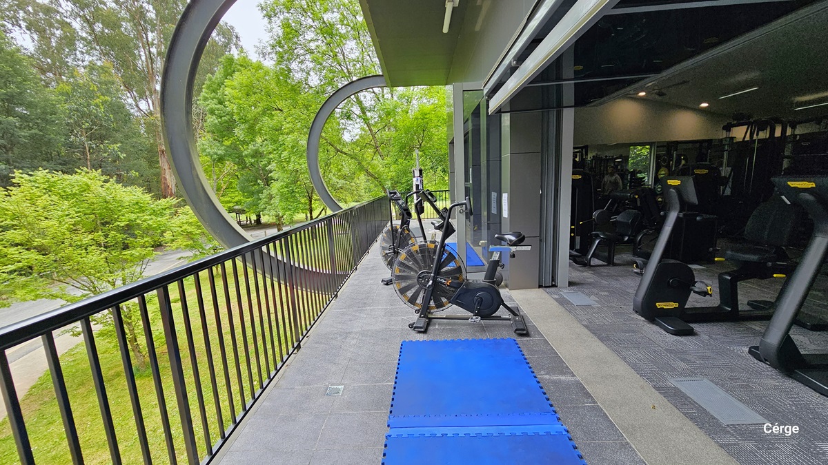 Facing the right side of the Veranda in the Gymnasium at Monbulk Aquatic Centre. The veranda has black railings and a grey tiled floor with two blue rubber mats near a stationary bike. The outside wall is grey with circular metal wall extensions. Seen through the veranda are the trees and the beautiful landscape surrounding the facility. Facing the right side of the Veranda in the Gymnasium at Monbulk Aquatic Centre. The veranda has black railings and a grey tiled floor with two blue rubber mats near a stationary bike. The outside wall is grey with circular metal wall extensions. Seen through the veranda are the trees and the beautiful landscape surrounding the facility.