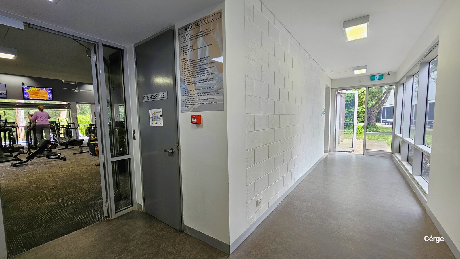 Corridor from the gymnasium leading to the exit glass doors. On the right wall from the gym entrance glass doors is a grey door with a FIRE HOSE REEL sign and a sign detailing the Health Club Rules. The floor is natural stone grey. The left wall is brick white, while the right is a combination of glass windows and white concrete wall. On the white ceiling are square fluorescent lights with warm light. Corridor from the gymnasium leading to the exit glass doors. On the right wall from the gym entrance glass doors is a grey door with a FIRE HOSE REEL sign and a sign detailing the Health Club Rules. The floor is natural stone grey. The left wall is brick white, while the right is a combination of glass windows and white concrete wall. On the white ceiling are square fluorescent lights with warm light.