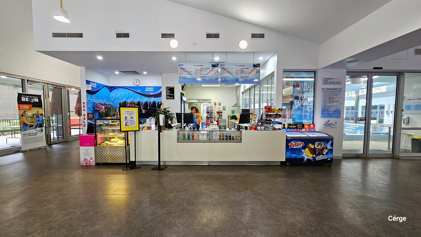Reception desk and café inside Monbulk Aquatic Centre. The reception desk is white with a middle glass display containing varied types, sizes, and colors of drinks. In front of the desk is a white automated sanitizer dispenser placed on a short, black metal post. In front of the glass display for pastries is a small yellow banner for the Cergé companion app. On the blue wall section behind the glass display are different sorts of swimwear. Reception desk and café inside Monbulk Aquatic Centre. The reception desk is white with a middle glass display containing varied types, sizes, and colors of drinks. In front of the desk is a white automated sanitizer dispenser placed on a short, black metal post. In front of the glass display for pastries is a small yellow banner for the Cergé companion app. On the blue wall section behind the glass display are different sorts of swimwear.
