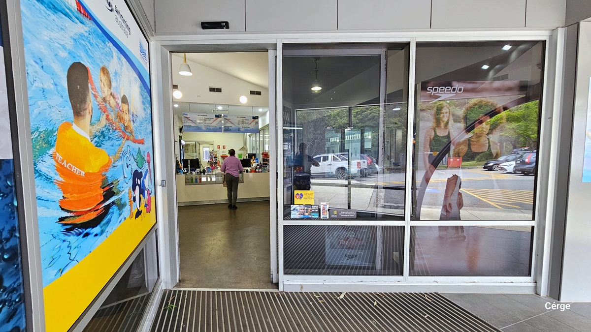 Automatic, glass sliding door entrance with white metal frames to Monbulk Aquatic Centre. On the left side of the entrance door is a large poster with an image of a male swimming coach wearing an orange shirt with the word TEACHER at the back of the shirt in white, guiding two children in the pool. Seen through the open door is a woman standing in front of a reception desk. The outside walls are gray, and the entrance floor has a stainless steel grating, while inside, is a white wall. Automatic, glass sliding door entrance with white metal frames to Monbulk Aquatic Centre. On the left side of the entrance door is a large poster with an image of a male swimming coach wearing an orange shirt with the word TEACHER at the back of the shirt in white, guiding two children in the pool. Seen through the open door is a woman standing in front of a reception desk. The outside walls are gray, and the entrance floor has a stainless steel grating, while inside, is a white wall.