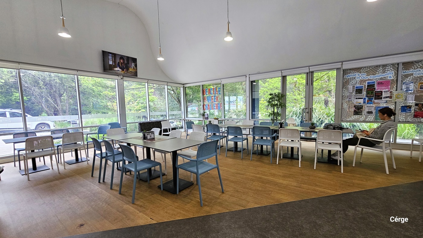 Seating area of the café. The floor space is a combination of brown and grey flooring. There are white square tables with black metal leg support and white and sky blue netted chairs. Near the right corner glass window is a red and blue ‘BE ACTIVE 6 WEEK CHALLENGE’ in capital letters. There is also a brown-and-white print bulletin board for What’s on in Our Community with photos and infographic materials. Mounted on the white wall, right above the glass window, is a flatscreen TV. Seating area of the café. The floor space is a combination of brown and grey flooring. There are white square tables with black metal leg support and white and sky blue netted chairs. Near the right corner glass window is a red and blue ‘BE ACTIVE 6 WEEK CHALLENGE’ in capital letters. There is also a brown-and-white print bulletin board for What’s on in Our Community with photos and infographic materials. Mounted on the white wall, right above the glass window, is a flatscreen TV.