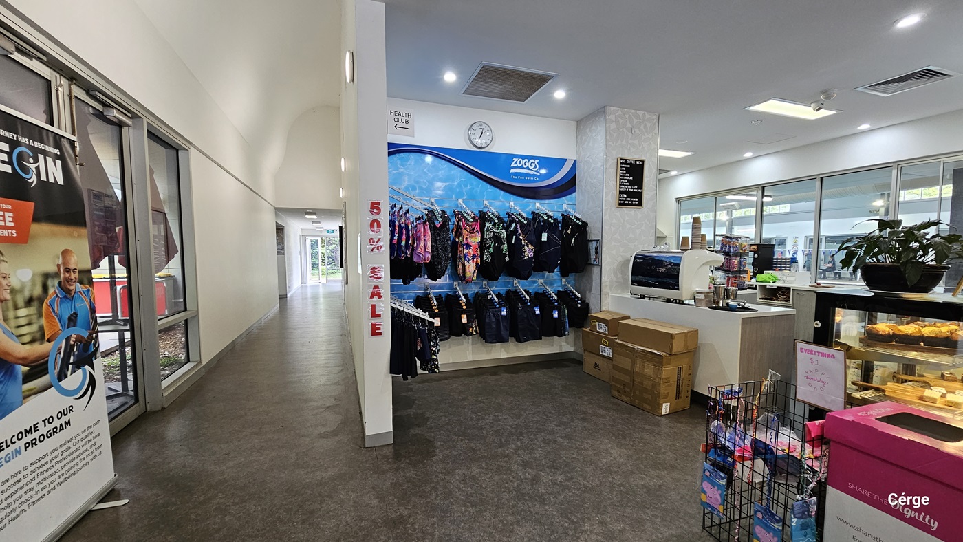 A closer look at the swimwear and swimming accessories of the reception desk and café inside Monbulk Aquatic Centre. The blue wall display at the back with the white ZOGGS logo showcases upper and lower swimwear for kids. Above it is a white clock, a white Health Club sign with an arrow pointing to the left passageway that has a large white gym advertisement at its entryway. Detailed on the white wall that separates the passageway and the café is a red 50% SALE. A closer look at the swimwear and swimming accessories of the reception desk and café inside Monbulk Aquatic Centre. The blue wall display at the back with the white ZOGGS logo showcases upper and lower swimwear for kids. Above it is a white clock, a white Health Club sign with an arrow pointing to the left passageway that has a large white gym advertisement at its entryway. Detailed on the white wall that separates the passageway and the café is a red 50% SALE.