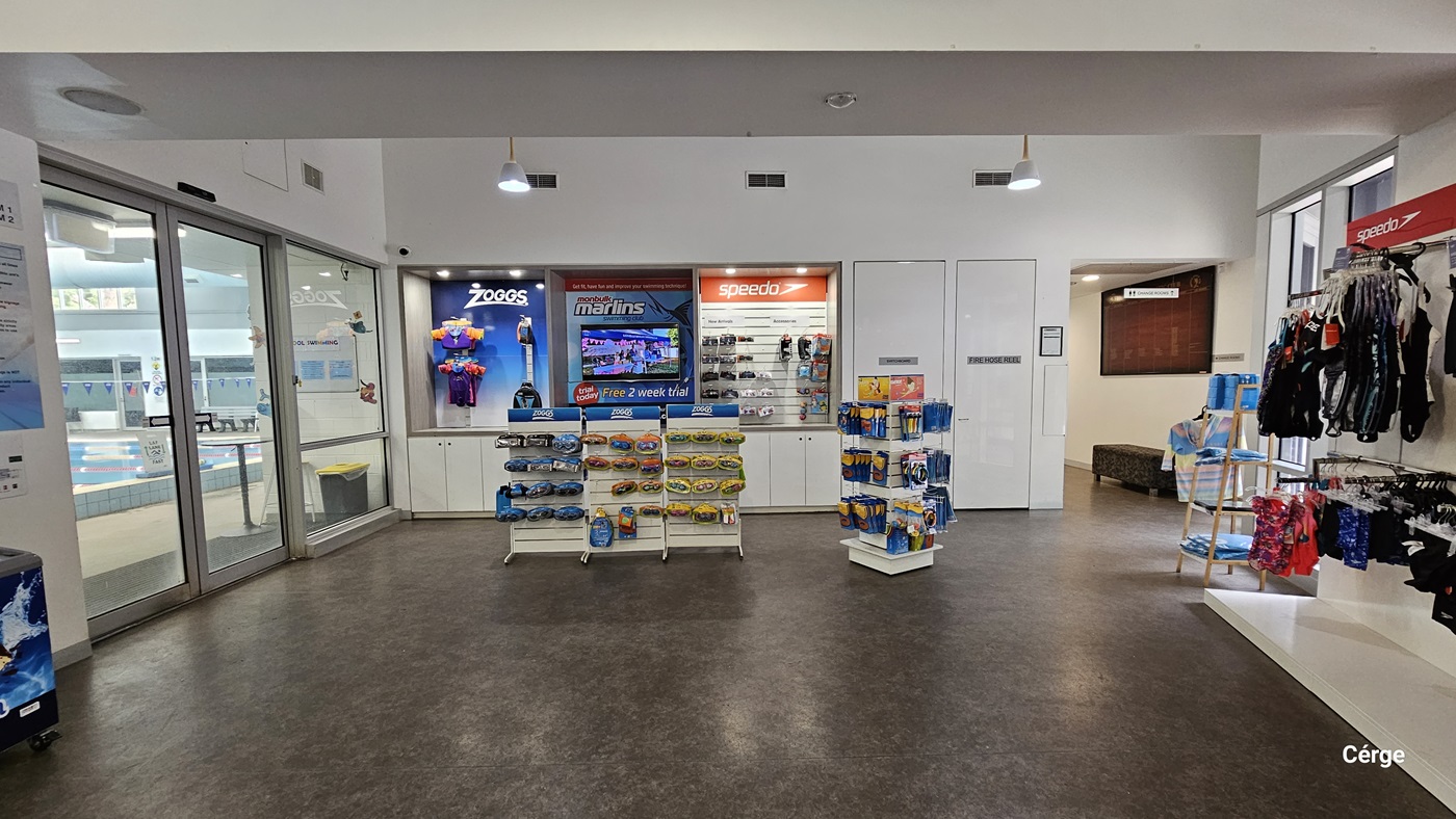 A display of swimwear and swimming accessories close to the entrance of the indoor pool accessible on the right of the reception and café. Mounted on the wall are upper and lower swimwear white on the white racks are swimming accessories such as goggles and swim caps. To the back are two white doors with a grey WHITE HOSE REEL sign next to a corner with a grey bench and a large, red glass wall display. The floor is natural stone grey, and the walls are white. A display of swimwear and swimming accessories close to the entrance of the indoor pool accessible on the right of the reception and café. Mounted on the wall are upper and lower swimwear white on the white racks are swimming accessories such as goggles and swim caps. To the back are two white doors with a grey WHITE HOSE REEL sign next to a corner with a grey bench and a large, red glass wall display. The floor is natural stone grey, and the walls are white.