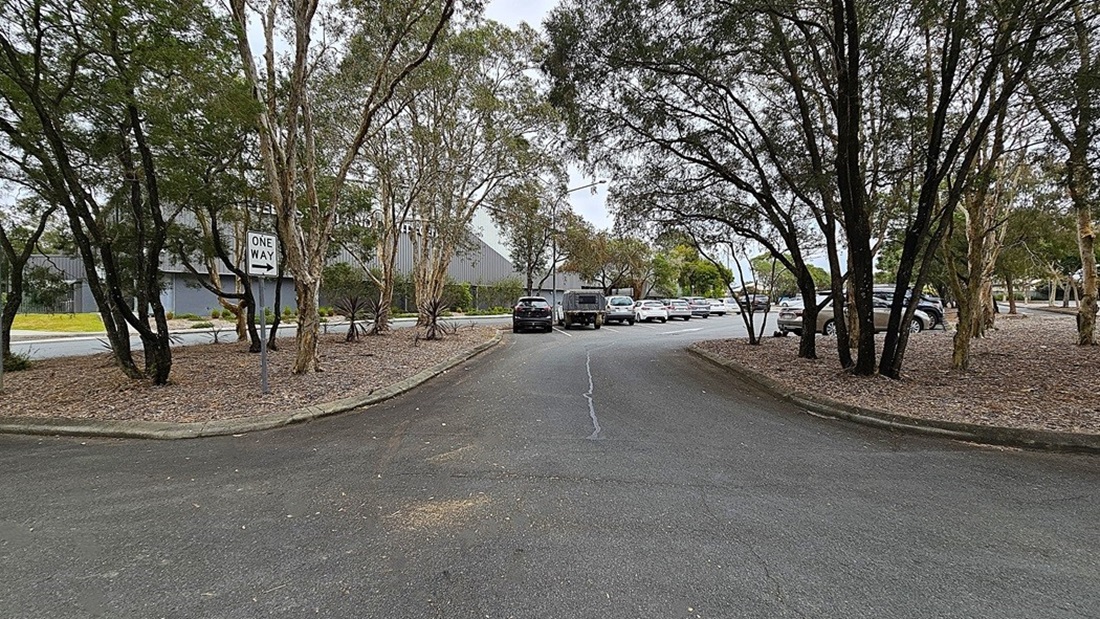 On the left, a row of trees lines the edge of the road, with a "ONE WAY" sign positioned among them. The ground is asphalt, showing a slight curve as it leads into a parking area. To the right, several parked cars are visible, with a mix of colours and sizes, and more trees provide a natural border. In the background, a large building with a grey exterior is partially obscured by the trees. The area is well-maintained, with gravel patches around the trees and clear pathways for vehicles.