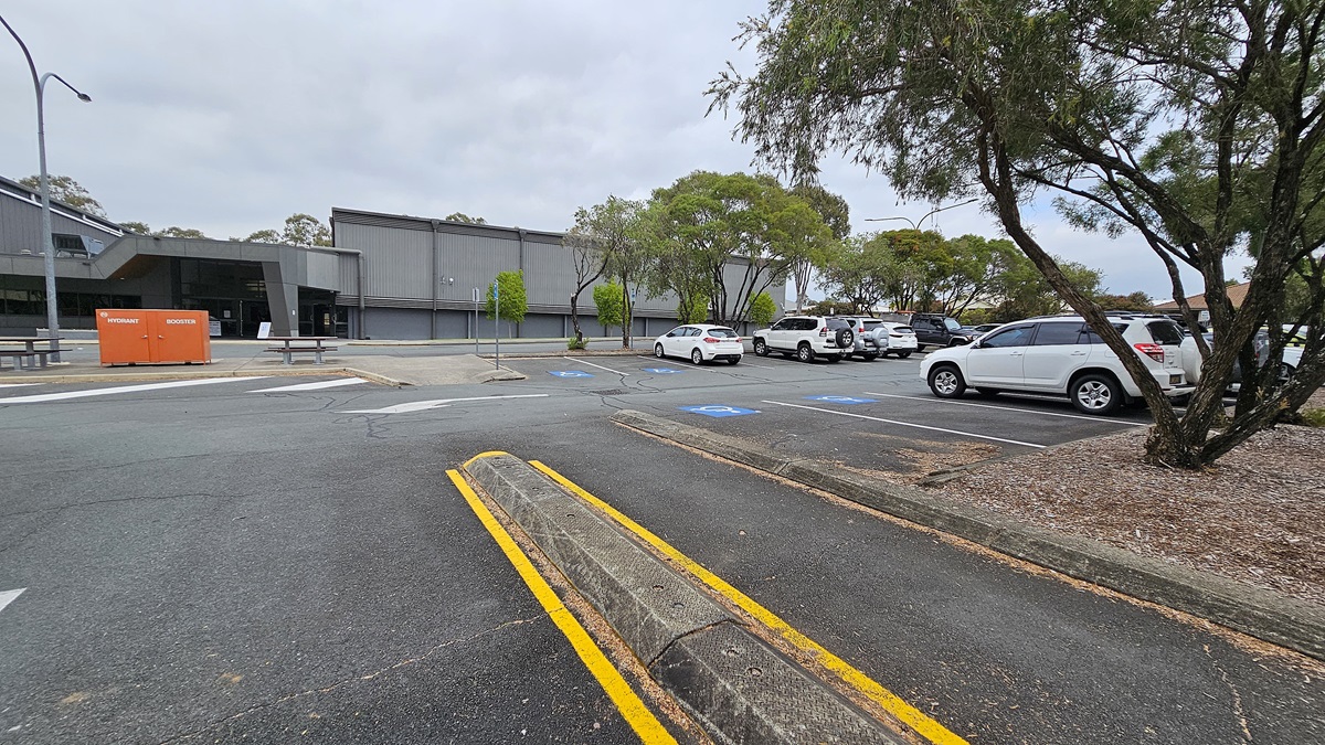On the left, there is a large grey building with a modern design, featuring a prominent entrance and a bright orange container labelled "RESTROOMS" positioned nearby. The ground is asphalt, with a slight incline leading towards the building. To the right, several parked cars are visible, predominantly white and silver, with a few trees providing some greenery. The parking area is well-defined, with marked spaces and a clean layout.