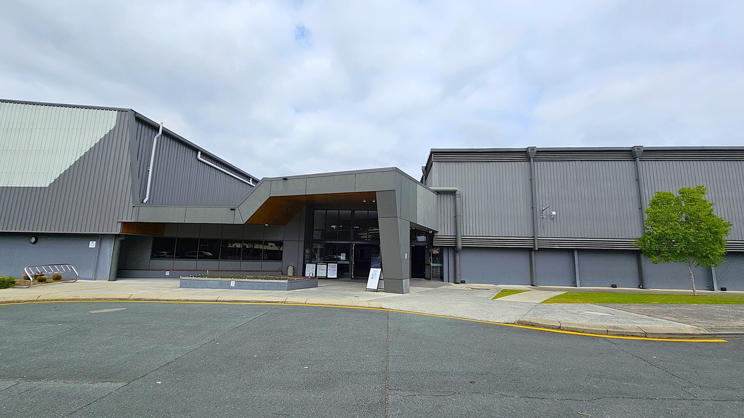 On the left, a large grey building features a sloped roof with vertical panels and a smooth facade. The entrance is highlighted by a modern overhang and large glass doors. A small landscaped area with low plants is visible in front of the entrance. To the right, the building continues with a similar grey exterior, and a small tree adds greenery. The ground is asphalt, with a curved pathway leading towards the entrance.