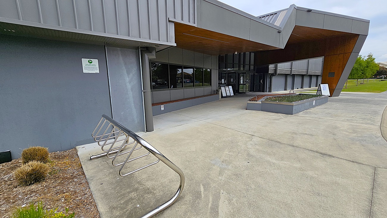 On the left, a grey wall features a sign and a bike rack with several slots, positioned next to a small patch of low shrubs. The ground is a smooth concrete surface, leading towards the entrance. In the centre, large glass doors are framed by a wooden overhang, providing a modern entrance. To the right, a raised planter box contains greenery, and the building continues with a similar grey facade.