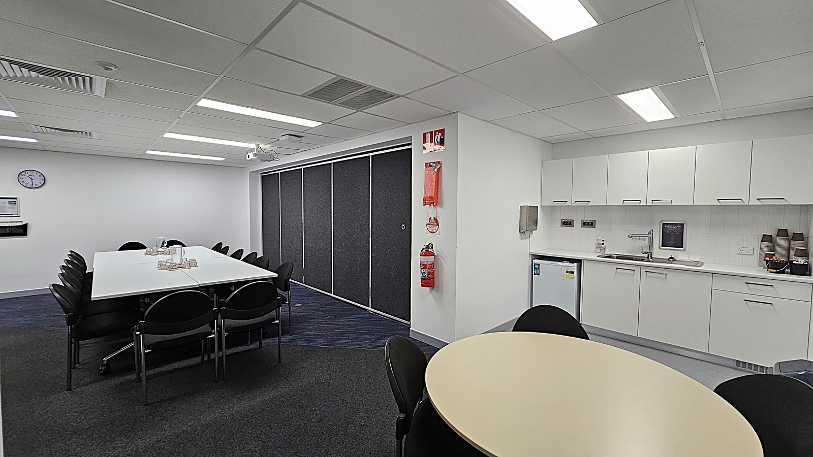 On the left, a long white table is surrounded by black chairs, with a glass pitcher and cups arranged on the table. The floor is dark grey carpet with blue accents. In the centre, a projector is mounted on the ceiling, aimed at a wall with grey blinds that can be drawn down. To the right, a black podium stands next to a whiteboard on a stand, with a few chairs positioned nearby. The ceiling is white with fluorescent lights.