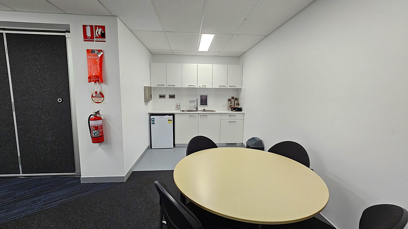 On the left, a sliding door leads to another room, while a wall features a fire extinguisher and safety signs. The floor is a combination of dark carpet and light grey tiles. In the centre, a round yellow table is surrounded by black chairs. To the right, a kitchenette includes white cabinets above a sink, with a small fridge below. A bin is positioned next to the fridge. The ceiling is white with fluorescent lighting.
