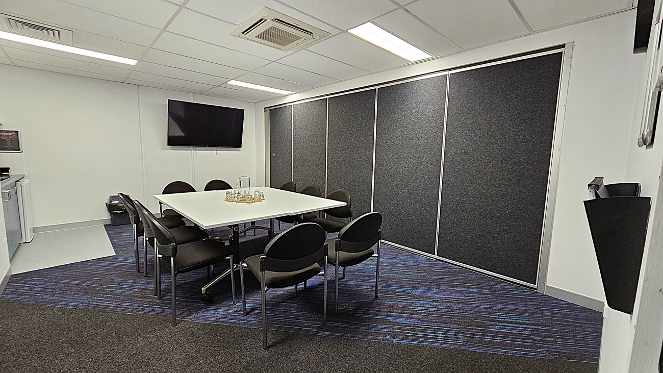 On the left, a kitchenette area features a white cabinet and a small sink, with a television mounted above. The floor is a mix of dark carpet and light grey tiles. In the centre, a white rectangular table is surrounded by black chairs, with glasses arranged on the table. To the right, a wall is covered with grey acoustic panels, and a sliding door is visible. The ceiling is white with fluorescent lights.
