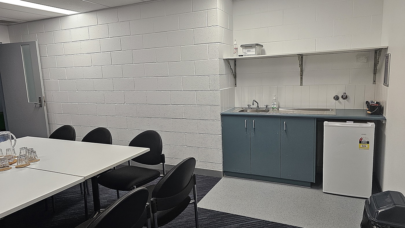 On the left, a grey door leads to another area, while a white wall features a smooth finish. The floor is a combination of dark carpet and light grey tiles. In the centre, a white rectangular table is surrounded by black chairs, with a glass pitcher and several glasses placed on the table. To the right, a kitchenette includes a dark blue cabinet with a sink and a small white fridge beneath it. Above, a shelf holds various items. The ceiling is white with fluorescent lighting.