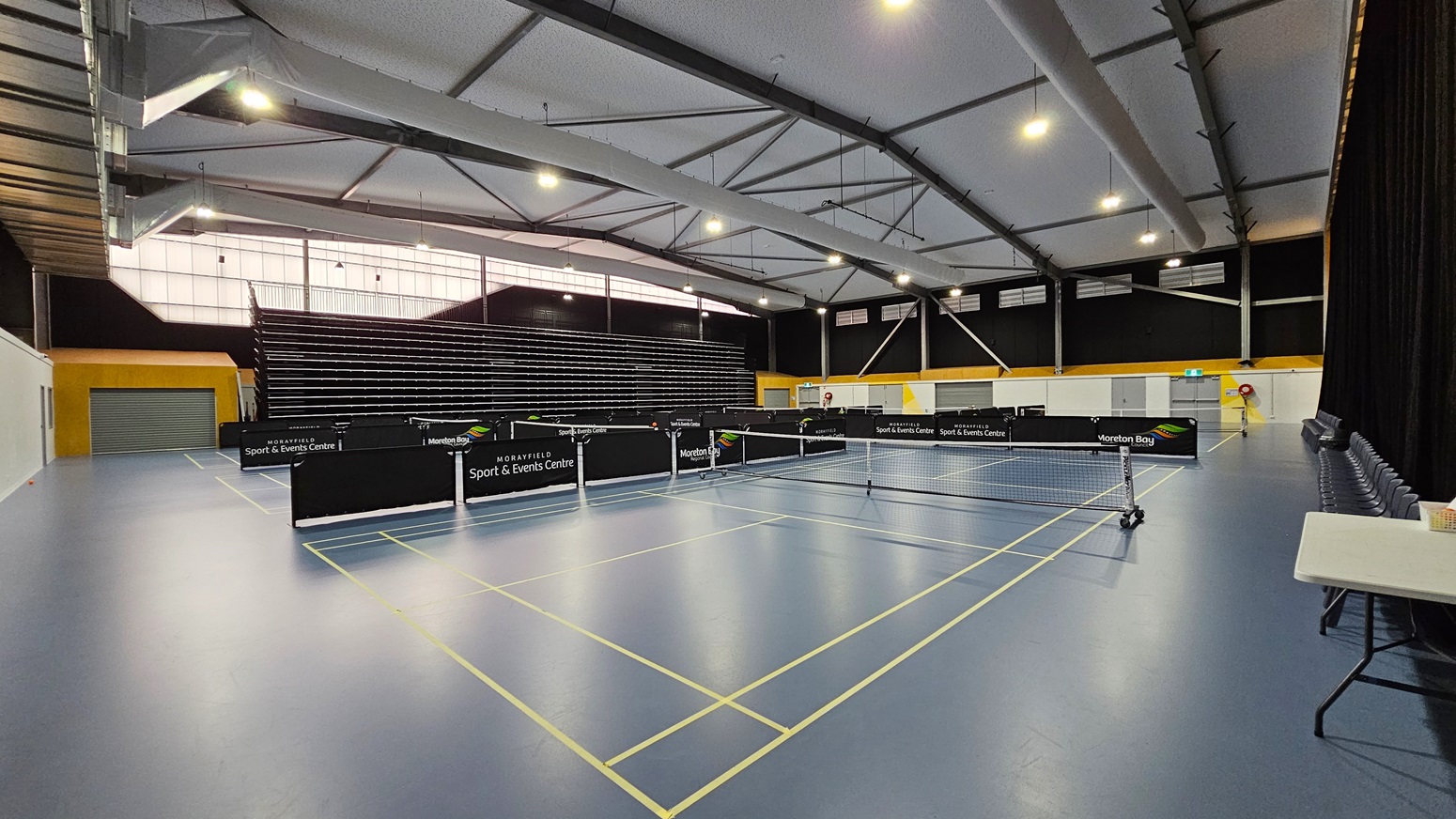 On the left, a service counter features a large screen displaying "COLLECT FOOD HERE," with a glass-fronted fridge filled with water bottles behind it. Two grey benches are positioned nearby. The floor is a mix of light blue and dark grey surfaces. In the centre, an open doorway leads to a large sports hall, where tables and a net are visible. To the right, the wall is painted blue, and the ceiling has exposed beams with spotlights.