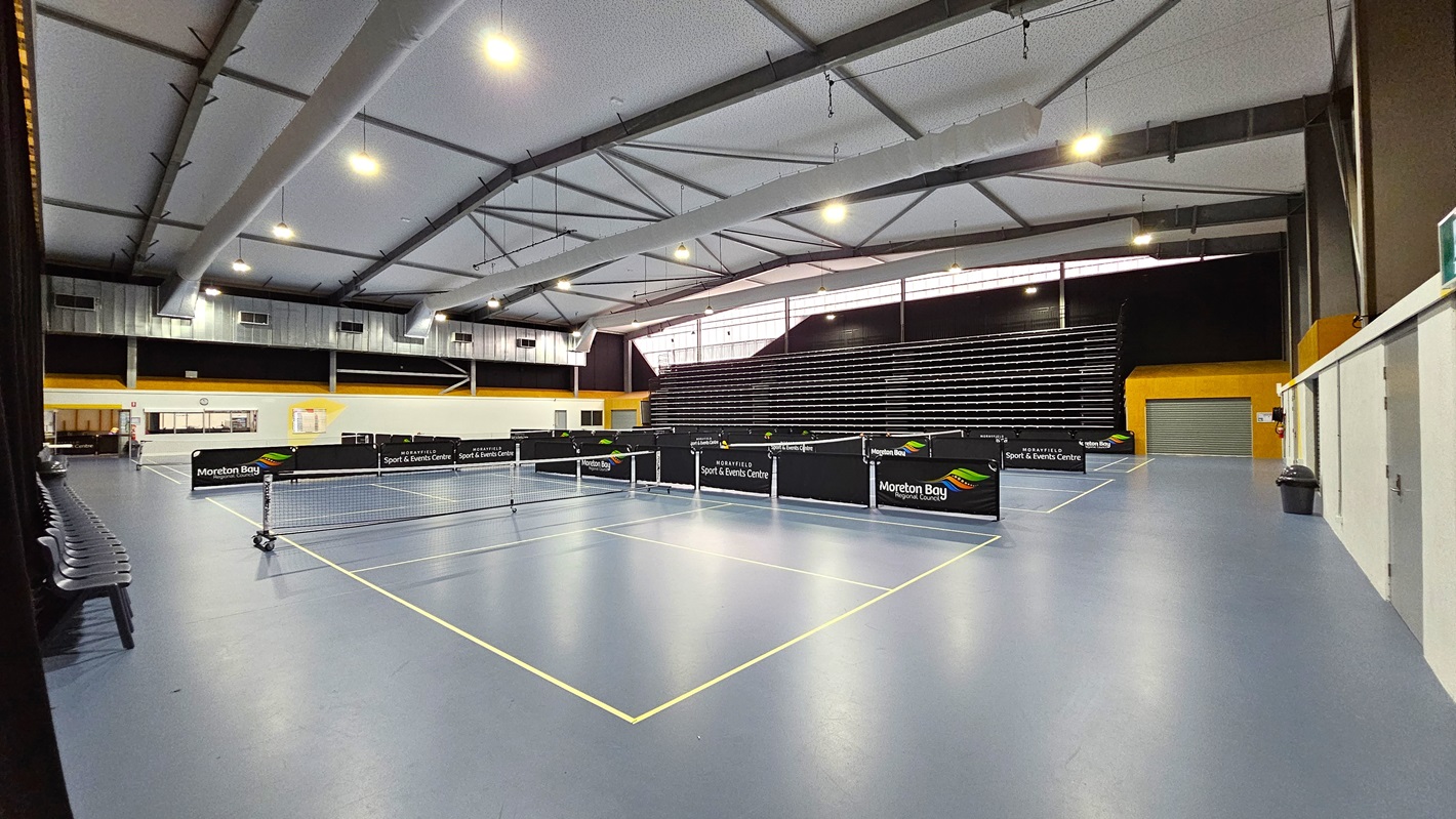 On the left, a row of black chairs is positioned against the wall, with a clock visible above. The floor is a smooth blue surface marked with white lines for table tennis. In the centre, several table tennis tables are arranged, each with a net in place. To the right, a wall features a yellow section and a closed grey door, with a bin nearby. The ceiling is high with exposed beams and bright lights. In the background, tiered seating is visible against a black wall.