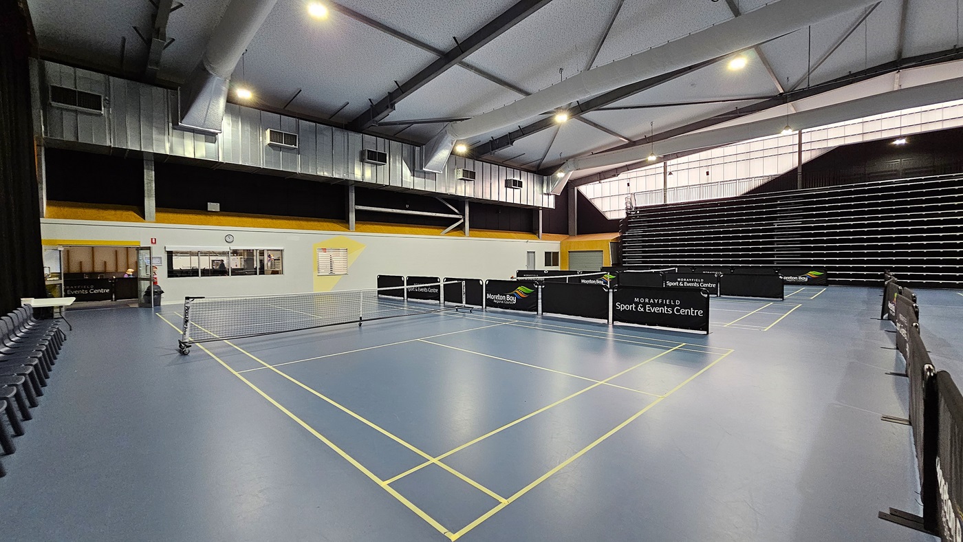 On the left, a row of black chairs is arranged against a white wall, with a clock mounted above. The floor is a smooth blue surface marked with yellow lines for table tennis. In the centre, several table tennis tables are set up, each with a net. To the right, a tiered seating area is visible, with black seating against a dark wall. The ceiling is high, featuring exposed beams and bright lights. A small service area is seen in the background, with a counter and windows.