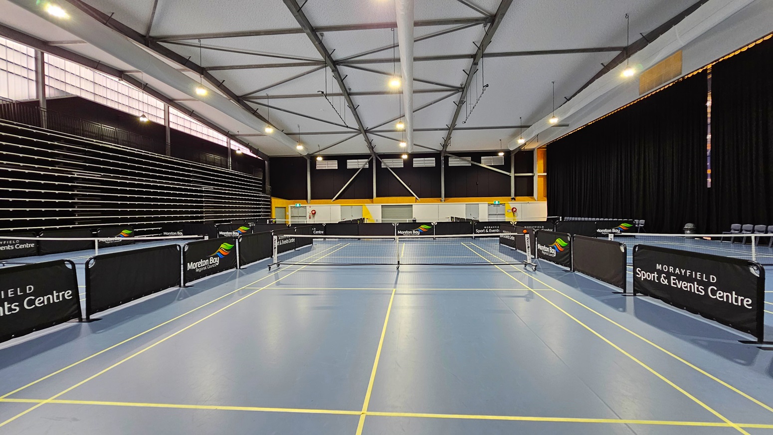 On the left, a tiered seating area is visible, with rows of black seats against a dark wall. The floor is a smooth blue surface marked with yellow lines for table tennis. In the centre, several table tennis tables are arranged, each with a net and surrounded by black barriers displaying the venue's name. To the right, a wall features a bright yellow section and a closed door, with fire extinguishers mounted nearby. The ceiling is high, with exposed beams and bright lights.